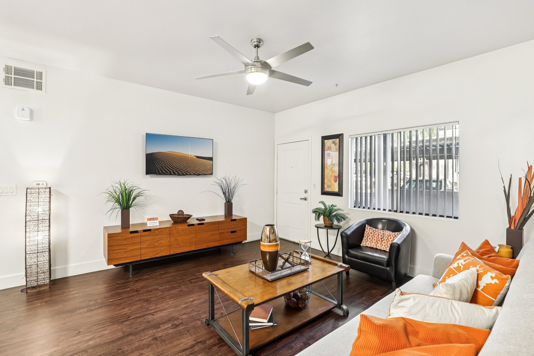 A modern living room featuring a light-colored sofa adorned with orange cushions, a wooden coffee table with decorative items, a sleek TV stand, and large windows with blinds. A ceiling fan circulates air, and potted plants add a touch of greenery, creating a warm and inviting atmosphere.