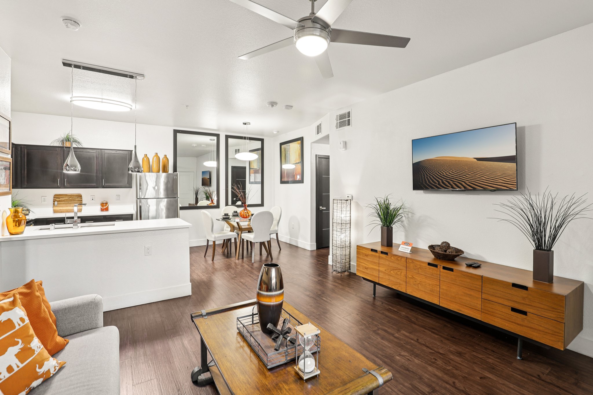 Modern living room and kitchen area featuring a sleek design. The space includes a comfortable sofa, a wooden coffee table, and a media console with a television. A dining table is visible in the background. Decorative plants and accents in warm colors enhance the inviting atmosphere, with natural light coming through large windows.