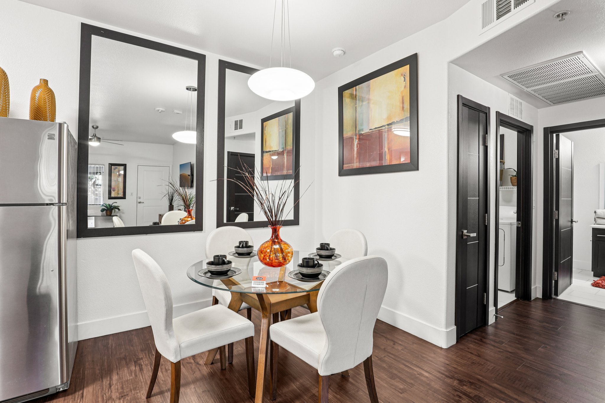 A modern dining area featuring a round glass table set with black dishes and a decorative orange vase. The walls have large mirrors and abstract paintings, while a stainless steel refrigerator is visible in the corner. The flooring is dark wood, creating a stylish and inviting atmosphere.