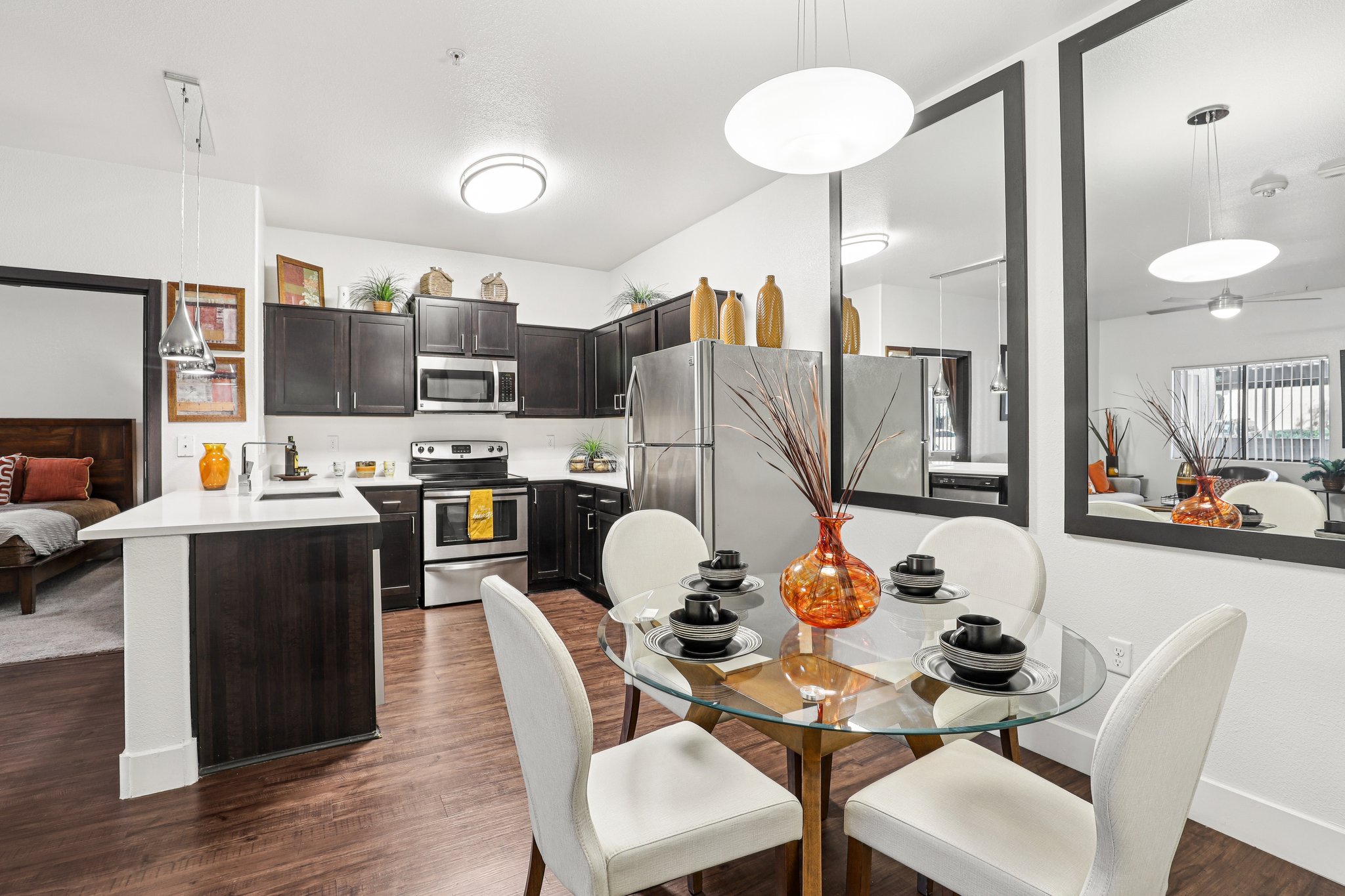 Modern kitchen and dining area featuring dark cabinetry, stainless steel appliances, and a glass dining table set for four. Decorative elements include a vase with twigs and orange accents. Large mirrors reflect light into the space, creating a bright and inviting atmosphere.