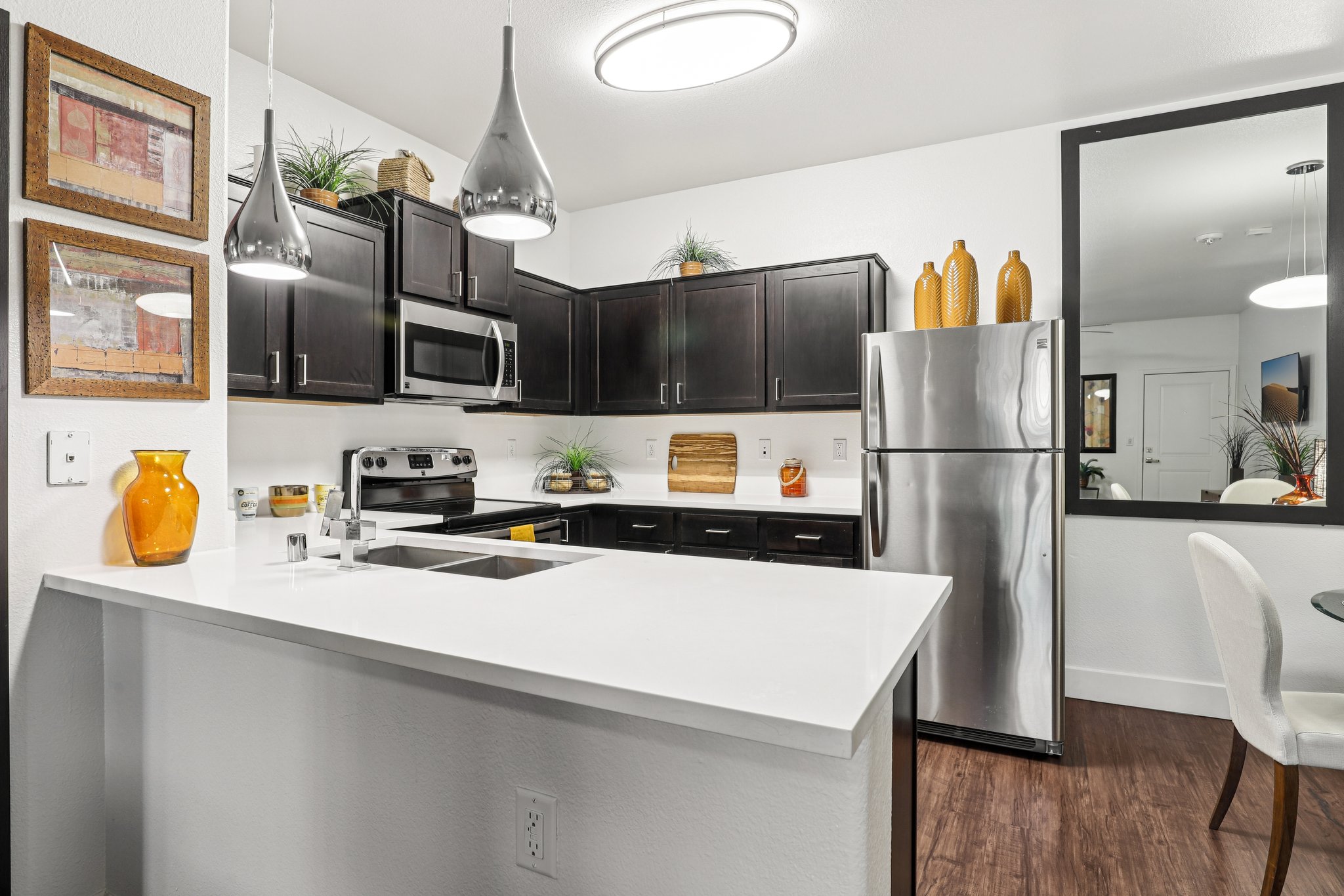 Modern kitchen featuring dark wooden cabinets, stainless steel appliances including a refrigerator and stove, a white countertop with a sink, pendant lighting, and decorative elements like potted plants and orange vases. The space has an open layout with a dining area visible in the background.