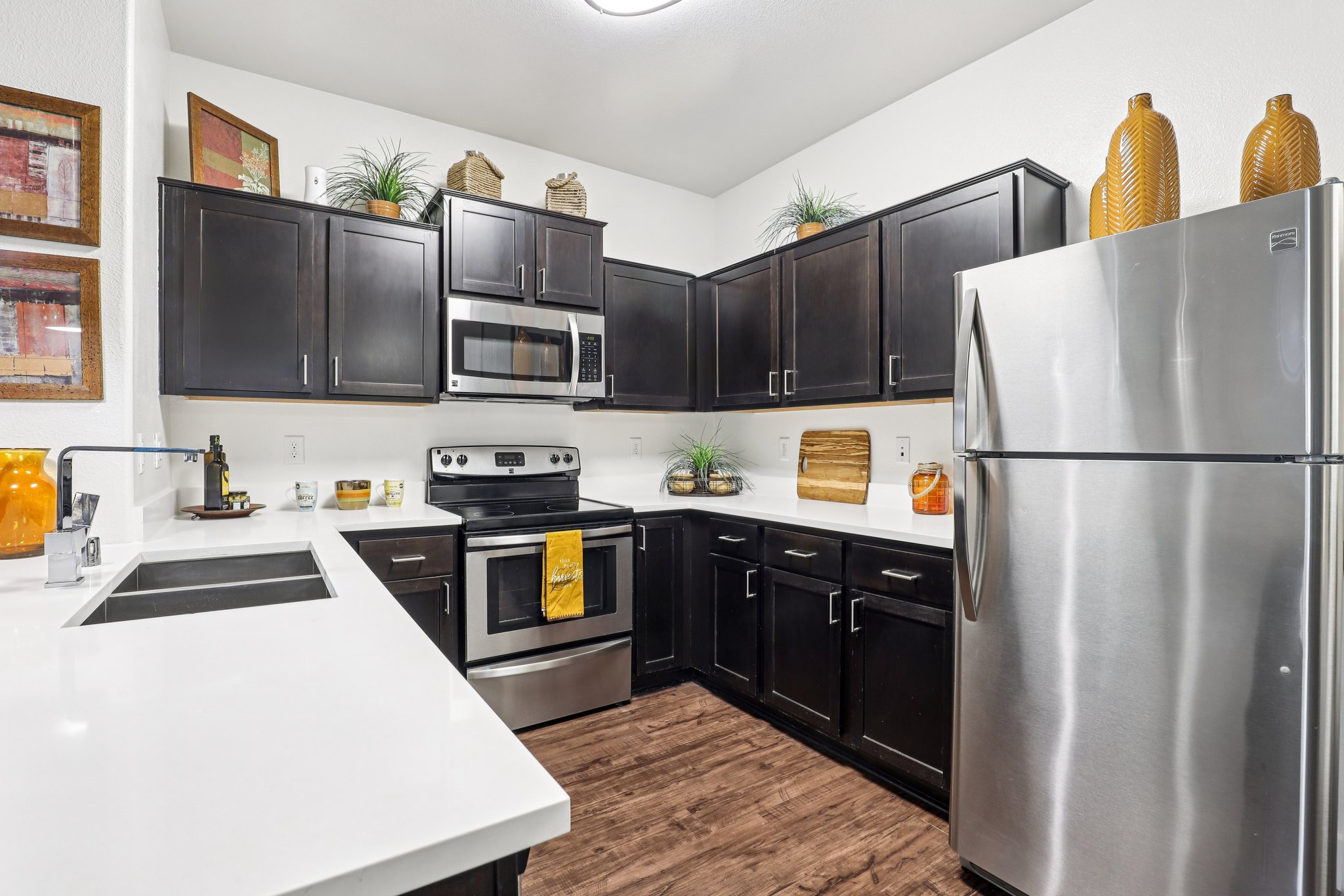 Modern kitchen featuring dark wood cabinets, stainless steel appliances, and a white countertop. Decor includes potted plants and decorative bowls. The space is bright and clean, with a functional layout highlighting the refrigerator and stove.