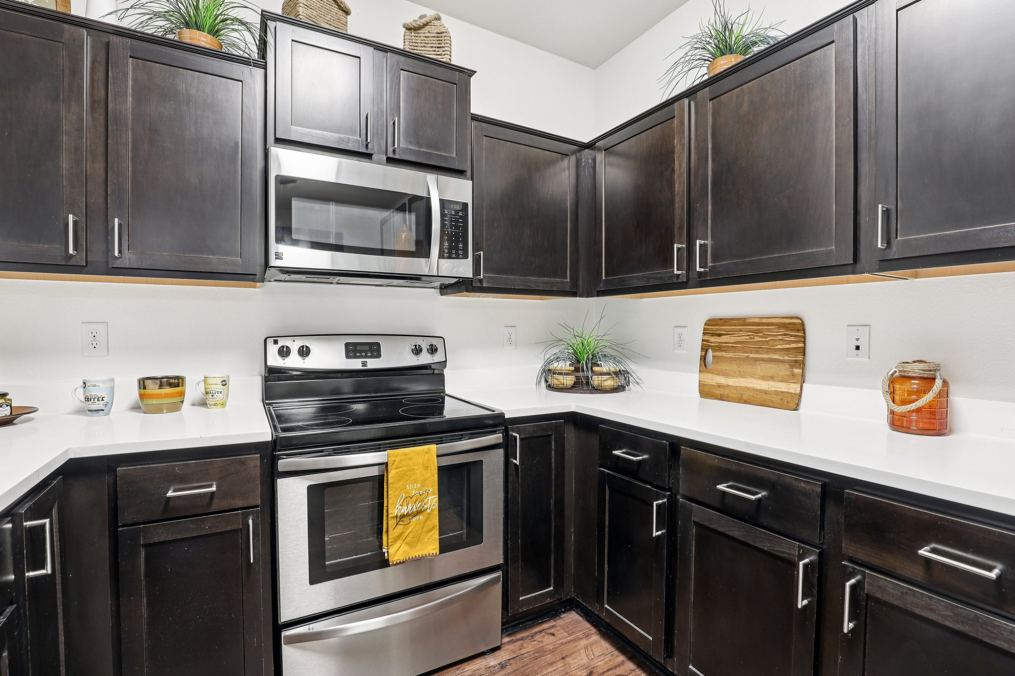 Modern kitchen featuring dark wooden cabinets, a stainless steel microwave and stove, white countertops, and decorative plants. A yellow kitchen towel hangs from the stove, and various kitchen accessories are displayed on the countertops, including a wooden cutting board and decorative bowls.