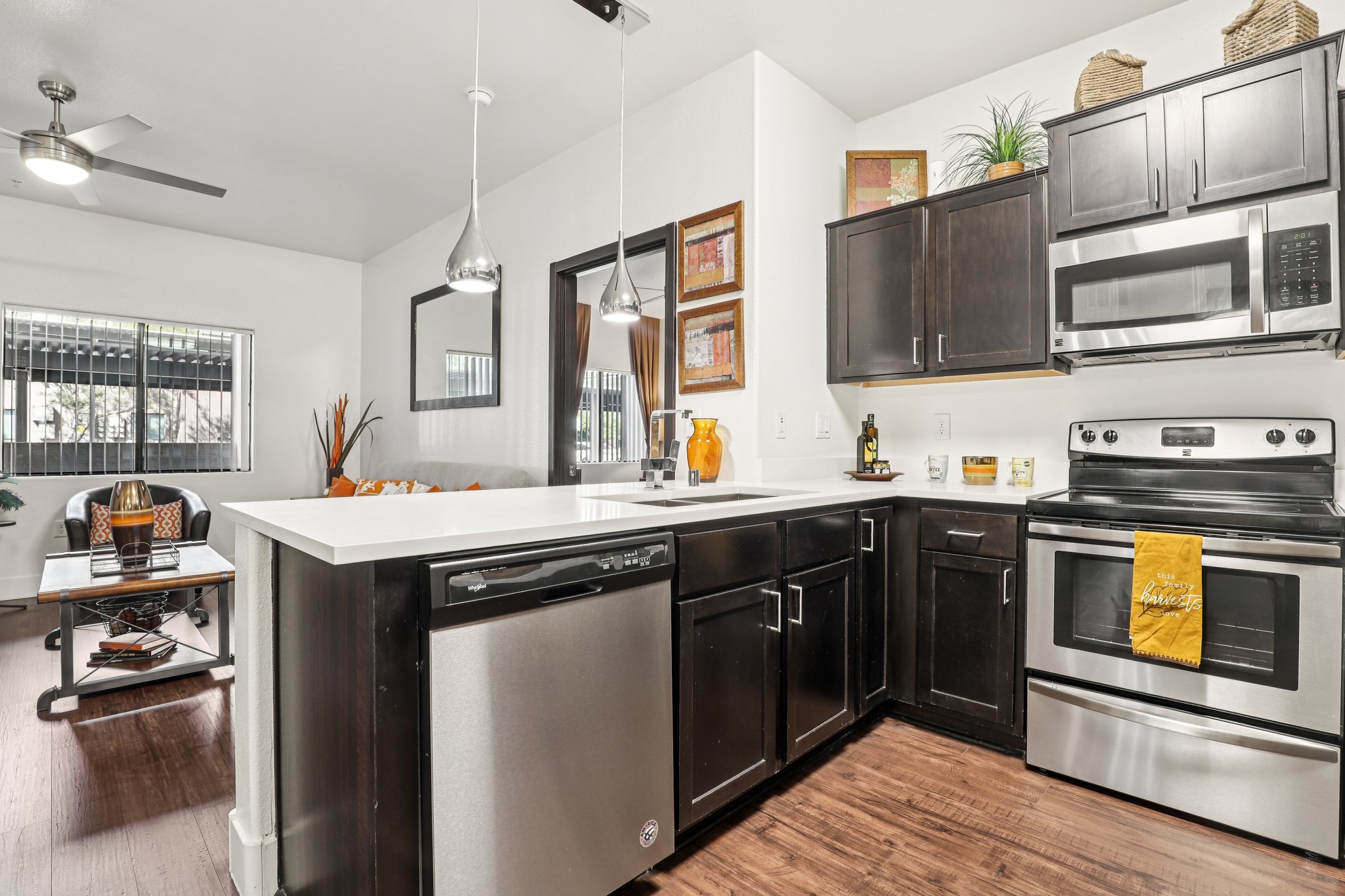 A modern kitchen featuring dark wood cabinets, stainless steel appliances, and a spacious countertop. A dishwasher is visible, alongside decorative items and a vibrant orange towel. Natural light streams in, highlighting a comfortable living area in the background with a table and sofa.