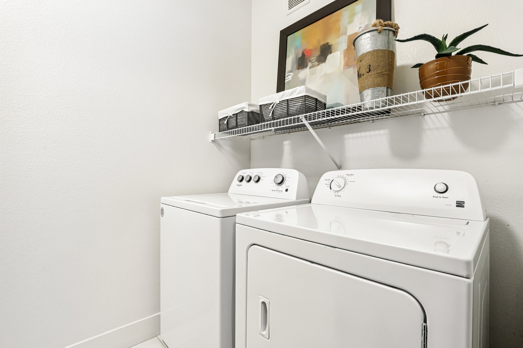 A clean laundry room featuring a white washing machine and dryer side by side. Above the appliances, there is a shelf holding neatly arranged storage baskets and a decorative plant. A framed abstract painting is mounted on the wall, adding a touch of color to the space.