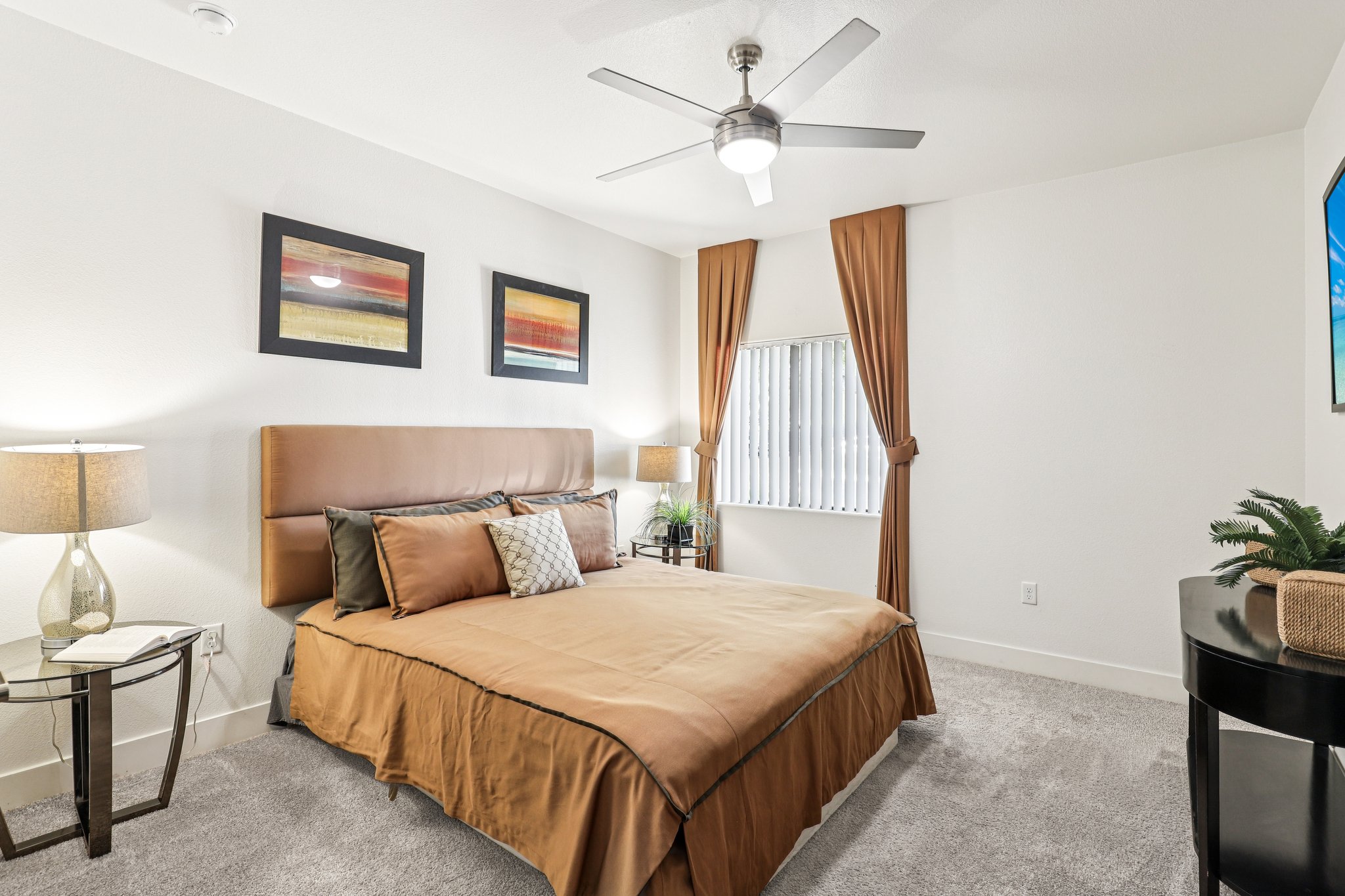 Modern bedroom featuring a large bed with a brown duvet, decorative pillows, and bedside lamps on each side. There are two framed artworks on the wall above the bed, and light drapes covering a window. A ceiling fan is installed above, and a small table with a plant is positioned beside the bed.