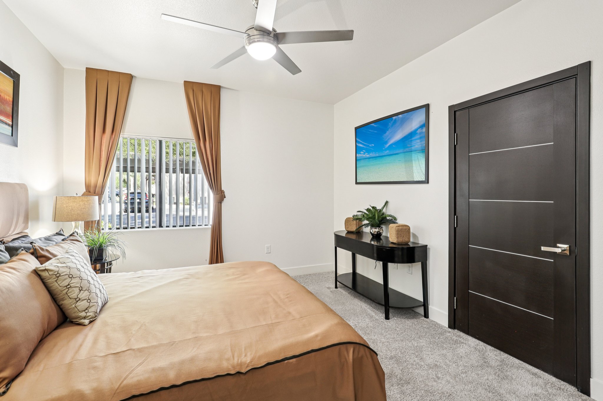 A well-lit bedroom featuring a large bed with a brown comforter and decorative pillows. There's a sleek black console table with plants and decorative items, a fan on the ceiling, and a large wall-mounted TV. Two curtains frame a window, adding warmth to the room's modern décor.