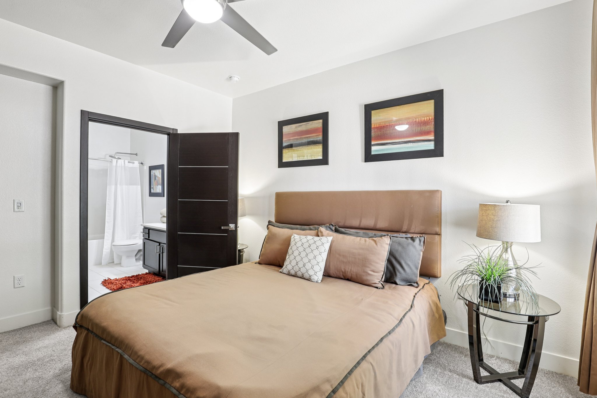 A cozy bedroom featuring a brown bedspread, decorative pillows, and modern artwork on the walls. There is a lamp on a small side table and a doorway leading to a bathroom with a shower curtain in the background. Soft lighting creates a warm and inviting atmosphere.