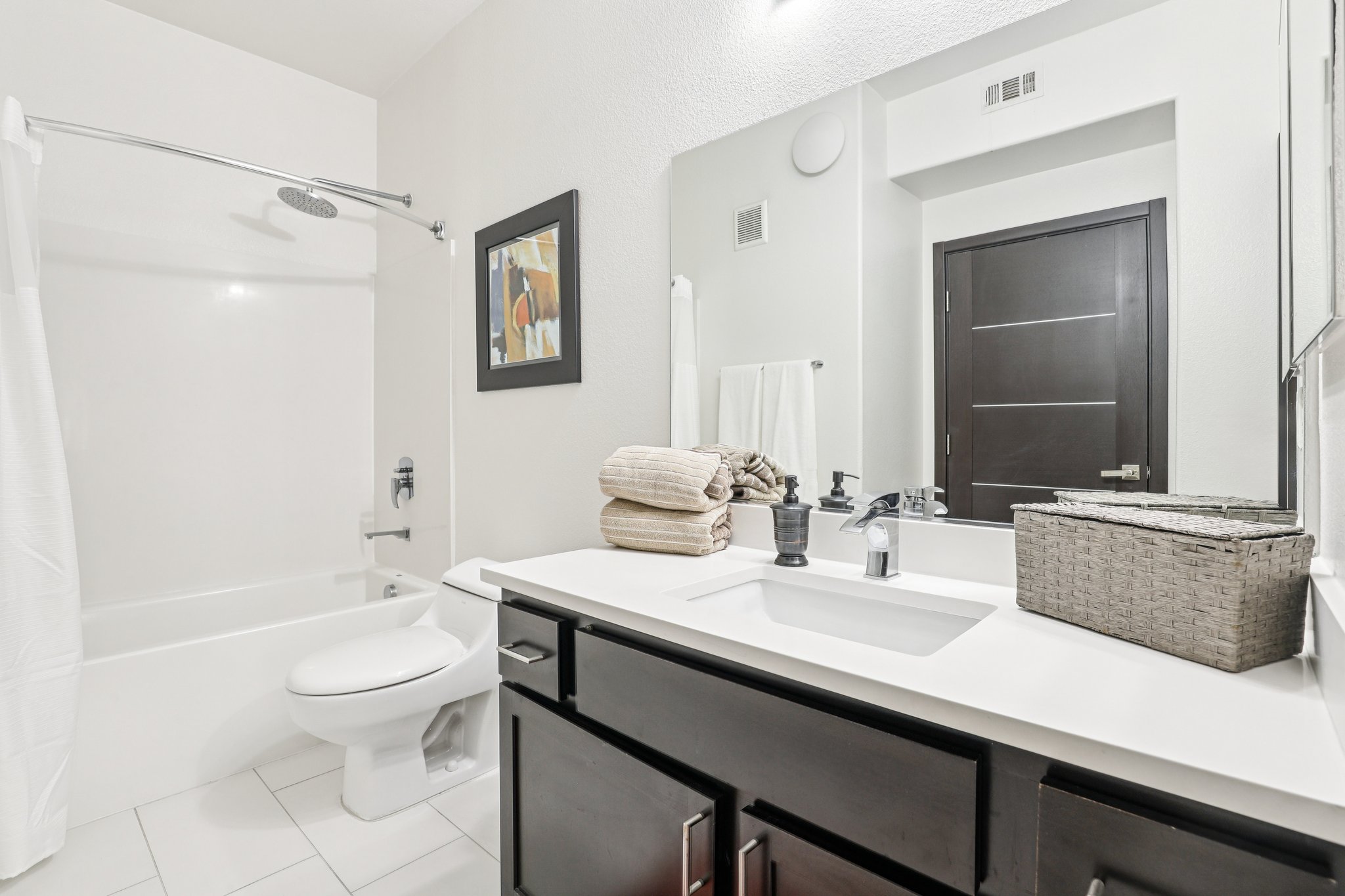 A modern bathroom featuring a white bathtub with a shower curtain, a dark wooden door, and a sleek vanity with a sink. The countertop is adorned with neatly stacked towels, a small plant, and a decorative basket. A mirror above the sink reflects the well-lit space.