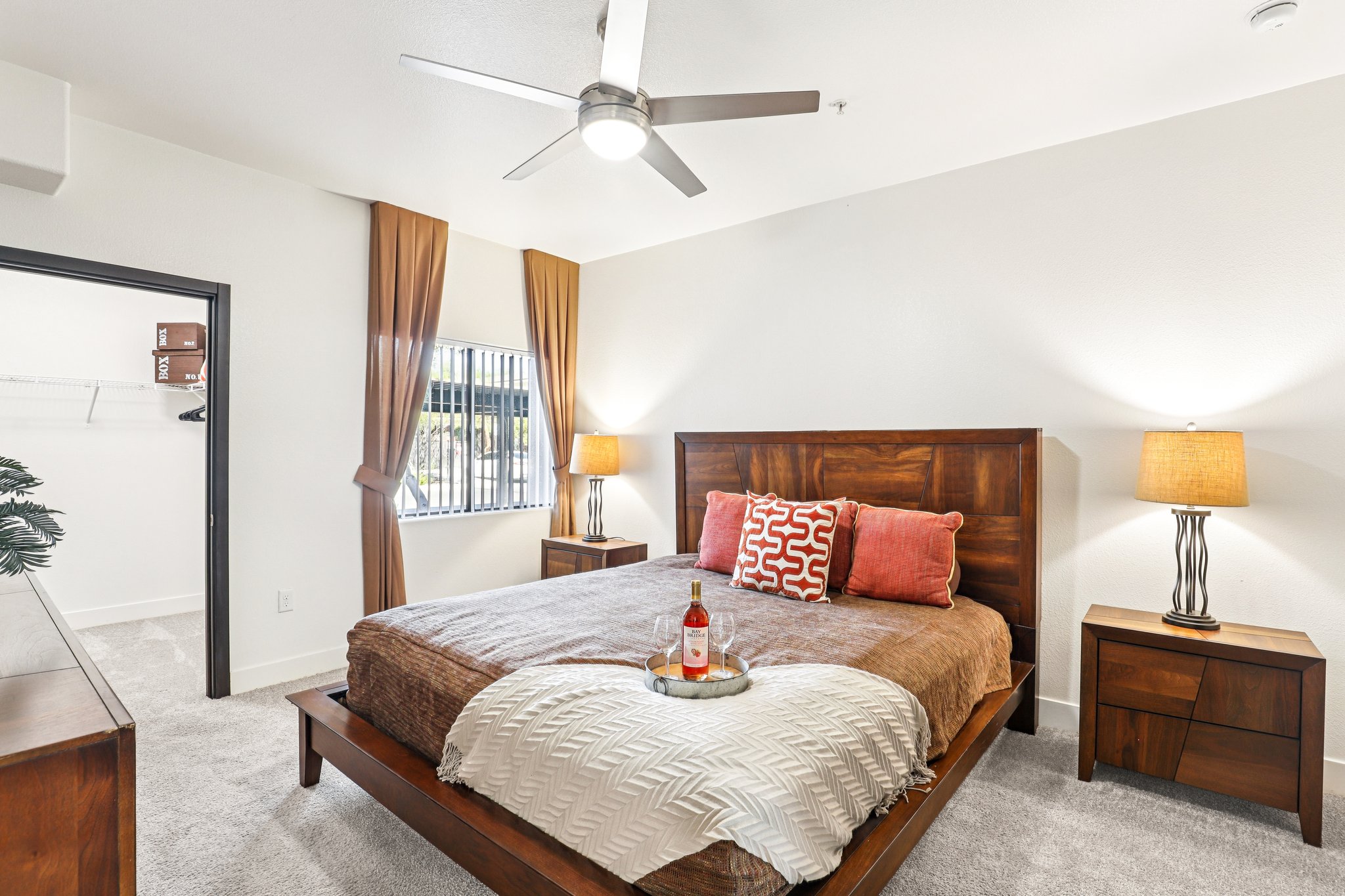 A cozy bedroom featuring a queen-sized bed with a wooden headboard, decorative pillows, and a throw blanket. Two bedside tables with lamps flank the bed. A large window with curtains adds natural light, and a ceiling fan is visible. The room has a neutral color scheme and carpeted flooring. A small tray with a bottle and glasses sits on the bed.