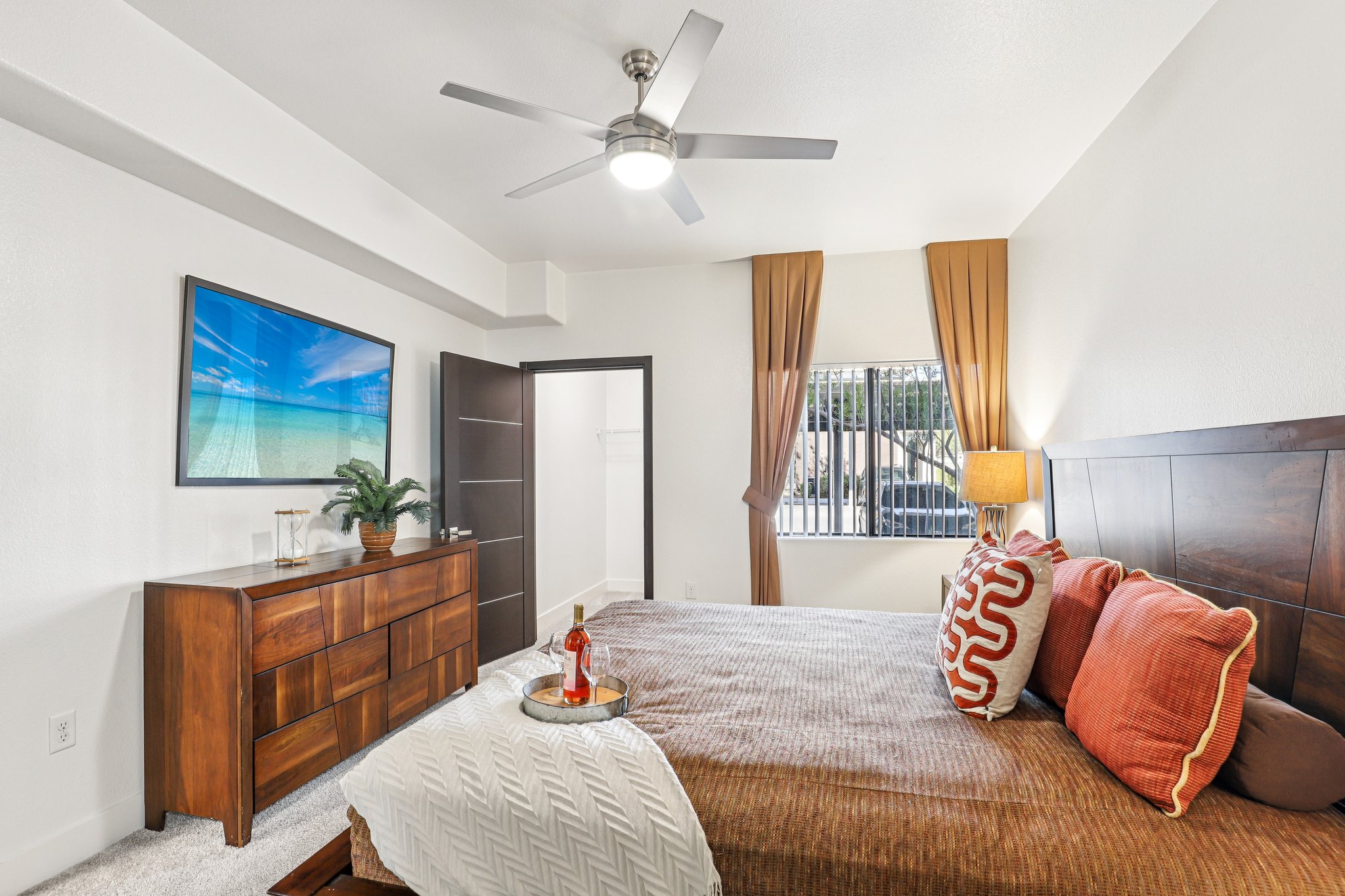 A modern bedroom featuring a queen-sized bed with orange and white patterned pillows, a wooden dresser, a ceiling fan, and a wall-mounted TV displaying a beach scene. Natural light comes through a window with gray curtains, and a decorative tray sits on the bed.
