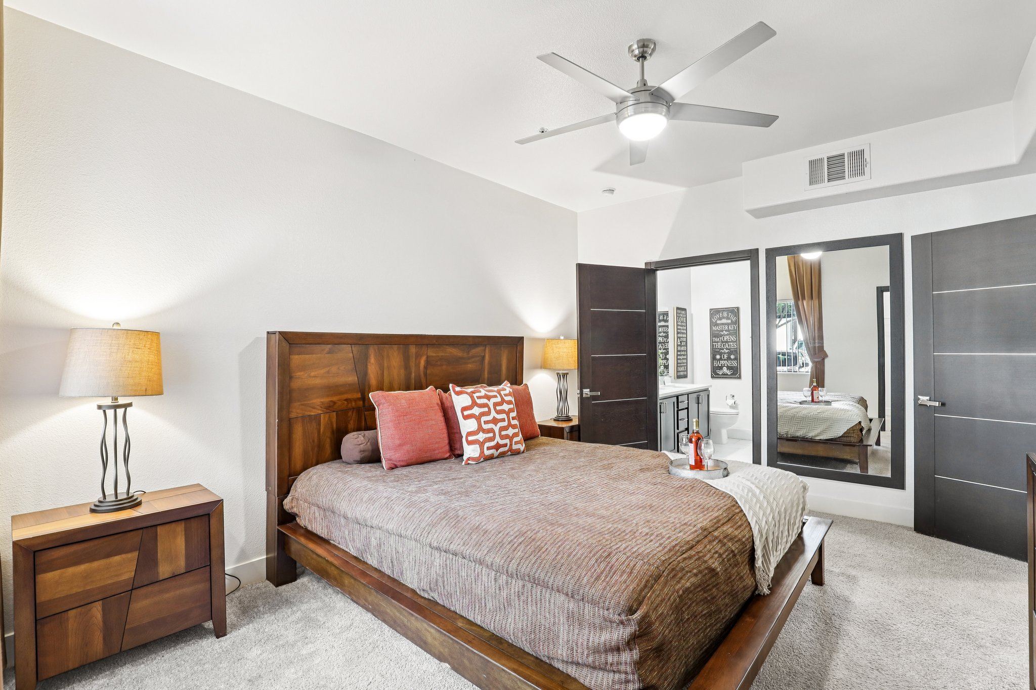 A stylish bedroom featuring a queen-sized wooden bed with decorative pillows, a nightstand with a lamp, and a large mirror reflecting a bathroom. The walls are light-colored, and the room includes soft carpeting and a ceiling fan, creating a cozy and inviting atmosphere.