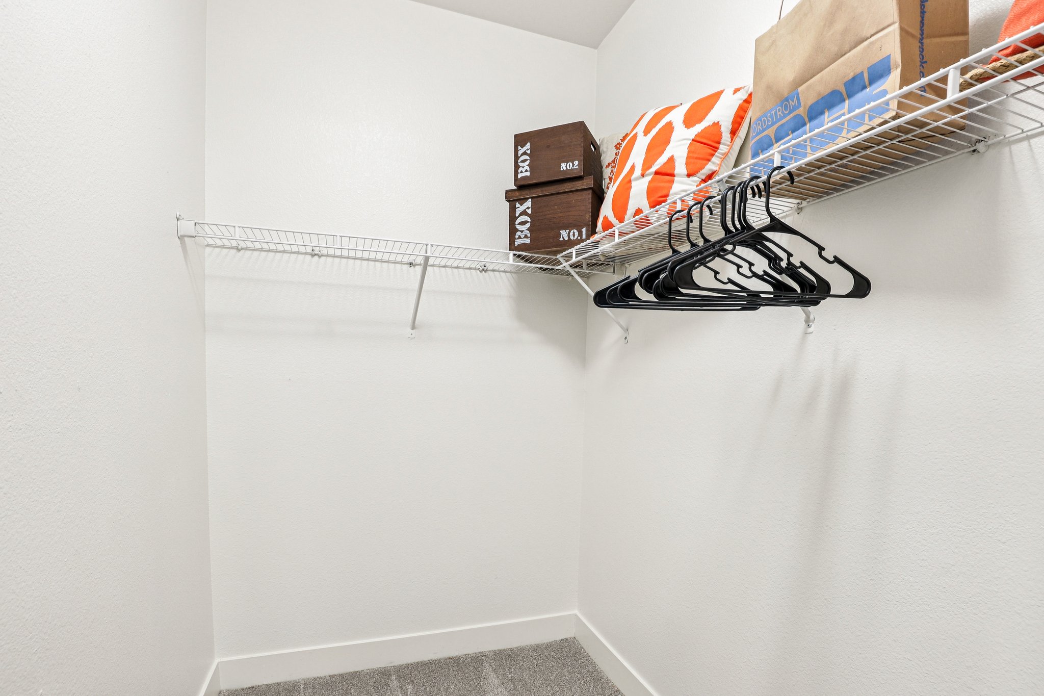 A tidy closet with a white wall and gray floor. It features a wire shelf holding two cardboard boxes and decorative pillows. Several black hangers are hanging from the shelf, with a portion of the closet empty, suggesting ample storage space.
