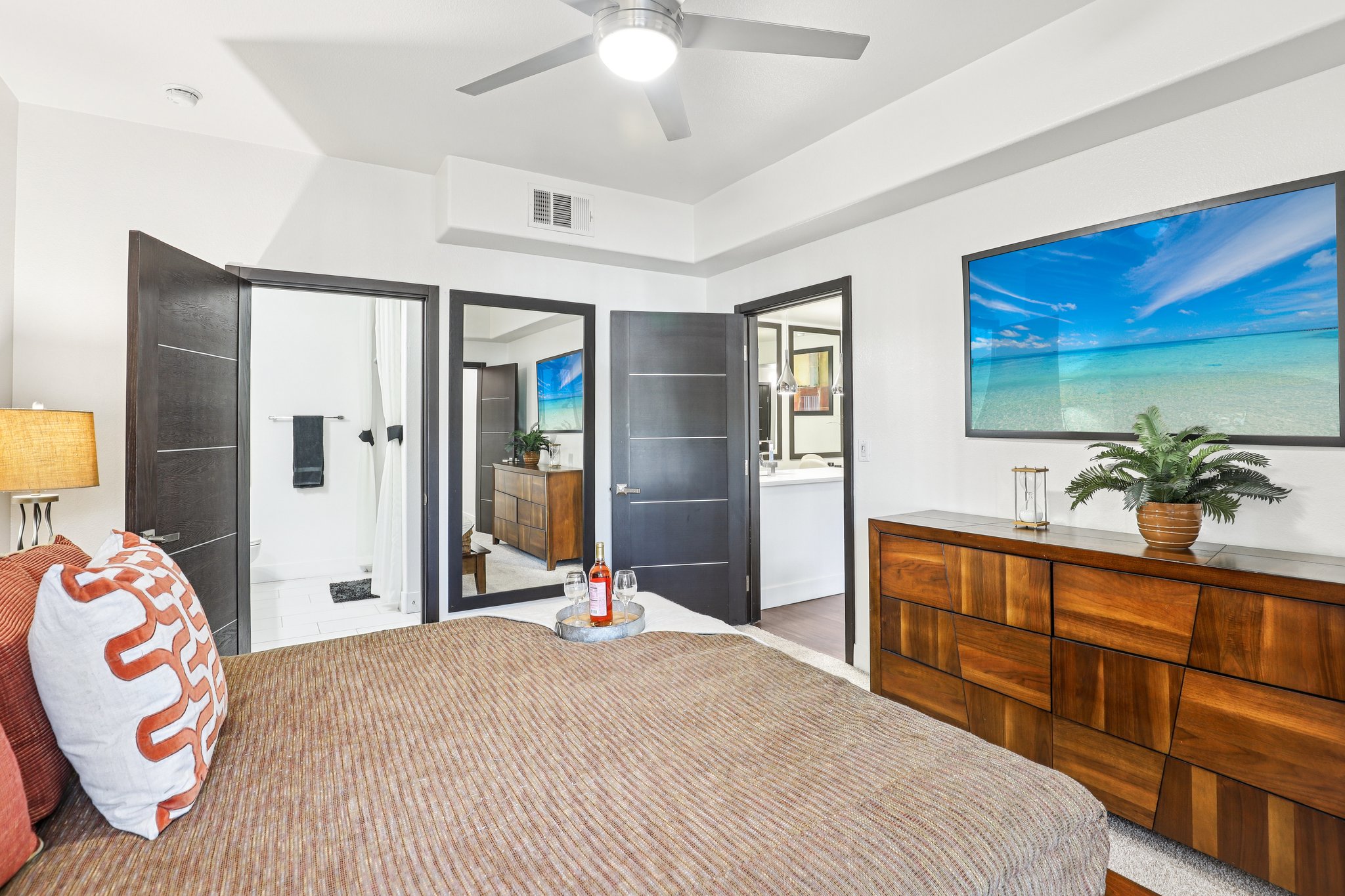 A modern bedroom featuring a bed with a decorative throw pillow and a wooden dresser. There is a ceiling fan above, and a large window with a beach scene artwork. Two doors lead to a bathroom, with one showing a shower area. The room has warm lighting and a potted plant on the dresser.