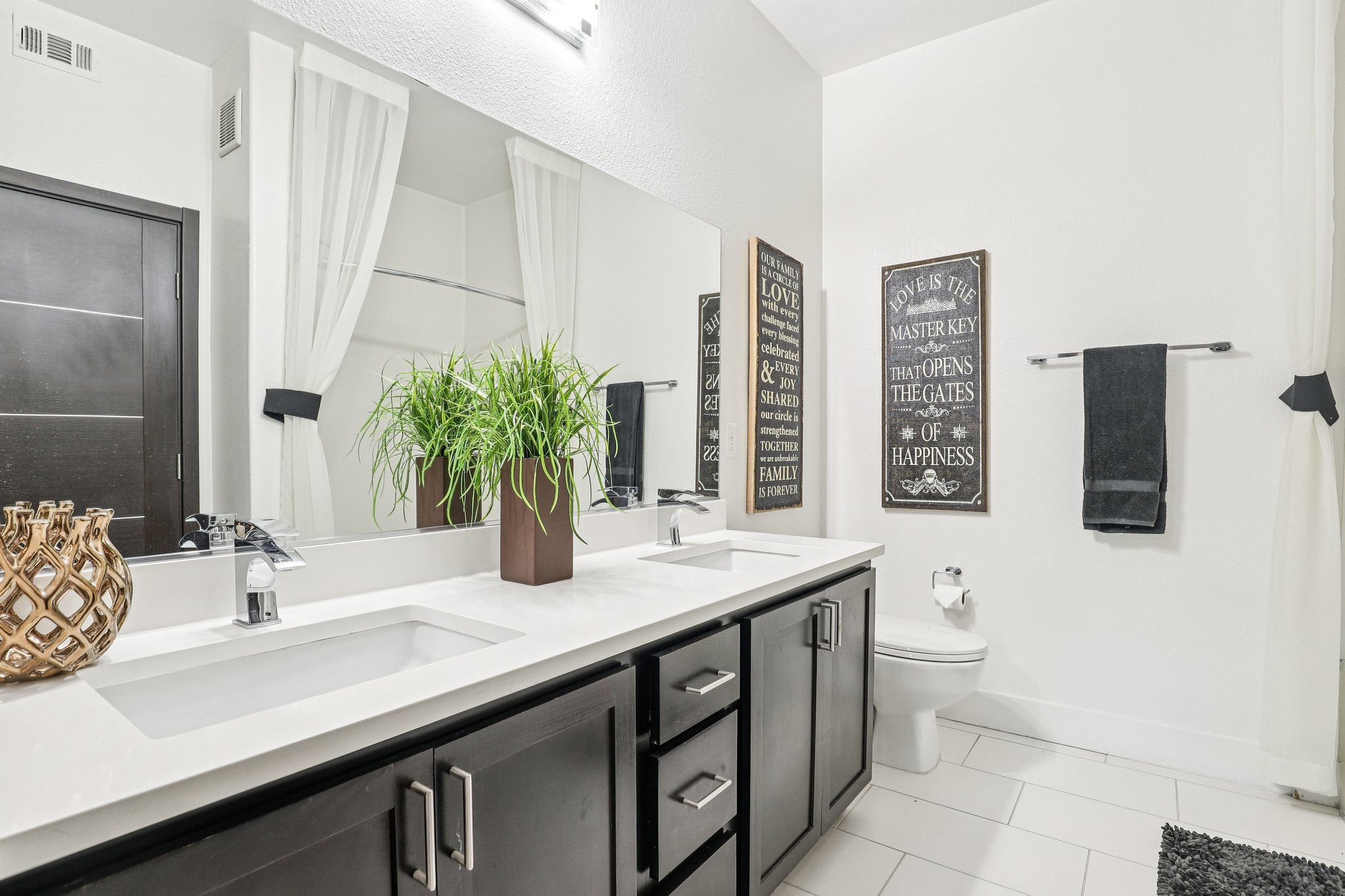 A modern bathroom featuring a double vanity with sinks, a large mirror, and decorative wall signs. The vanity has dark wood cabinets, a potted plant in a square brown pot, and a towel rack with a black towel. The decor is minimalistic with white walls and light tile flooring.