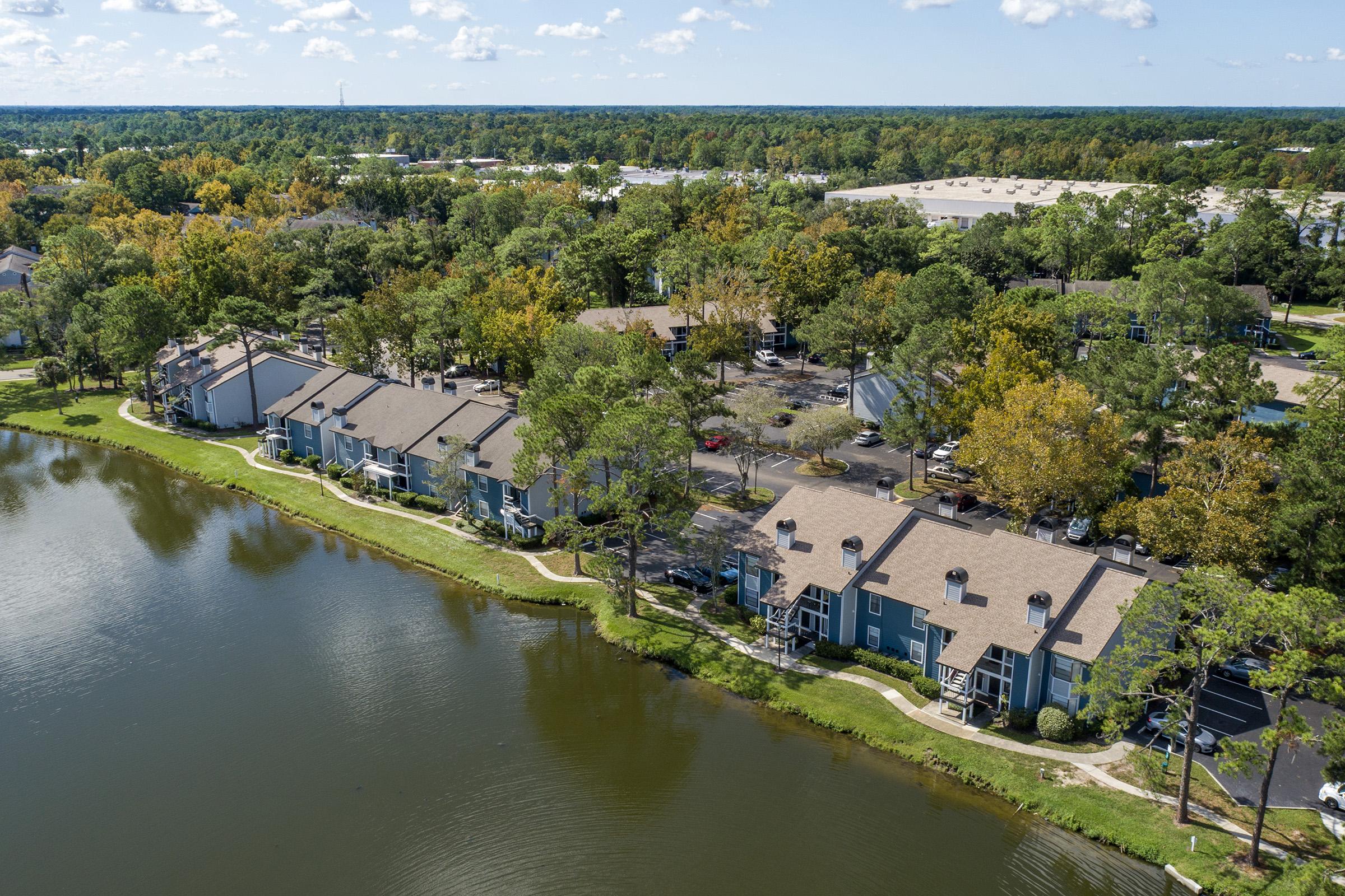 Aerial view of a residential area with two-story apartment buildings along a serene lake. Surrounding greenery features trees with autumn colors. The scene captures a peaceful suburban environment with blue skies and fluffy clouds above, showcasing a well-maintained community.