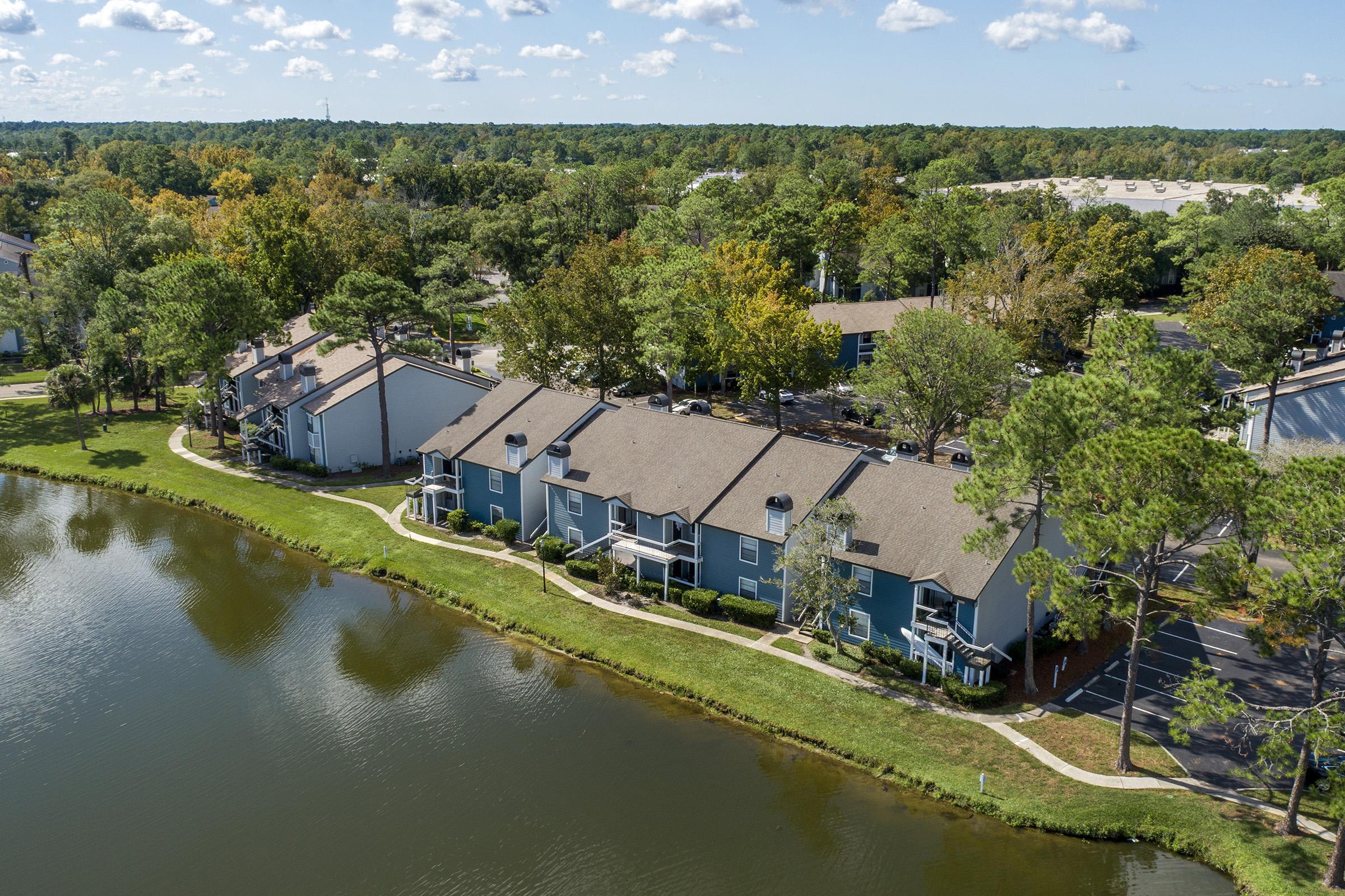 Aerial view of a residential area featuring multiple two-story apartment buildings along a serene pond. Lush greenery surrounds the buildings and pathways, with trees displaying autumn colors. The sky is partly cloudy, creating a tranquil suburban atmosphere.