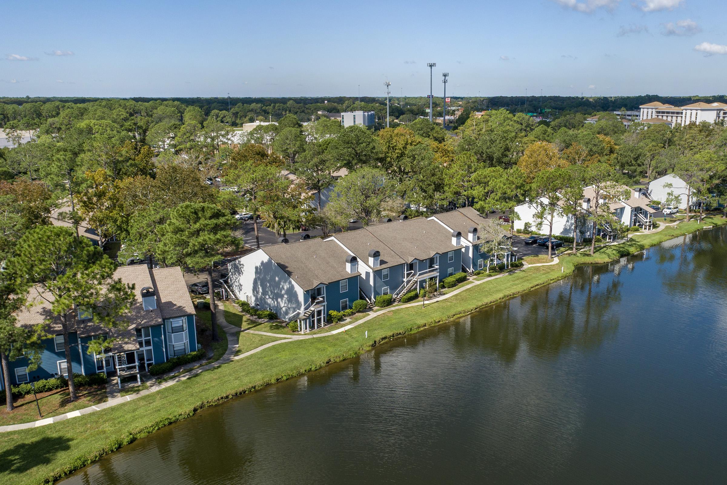 Aerial view of a residential area with two-story apartment buildings lined along a peaceful lake. The surrounding landscape features trees with autumn foliage. In the background, a mix of buildings and green spaces can be seen under a partly cloudy sky.