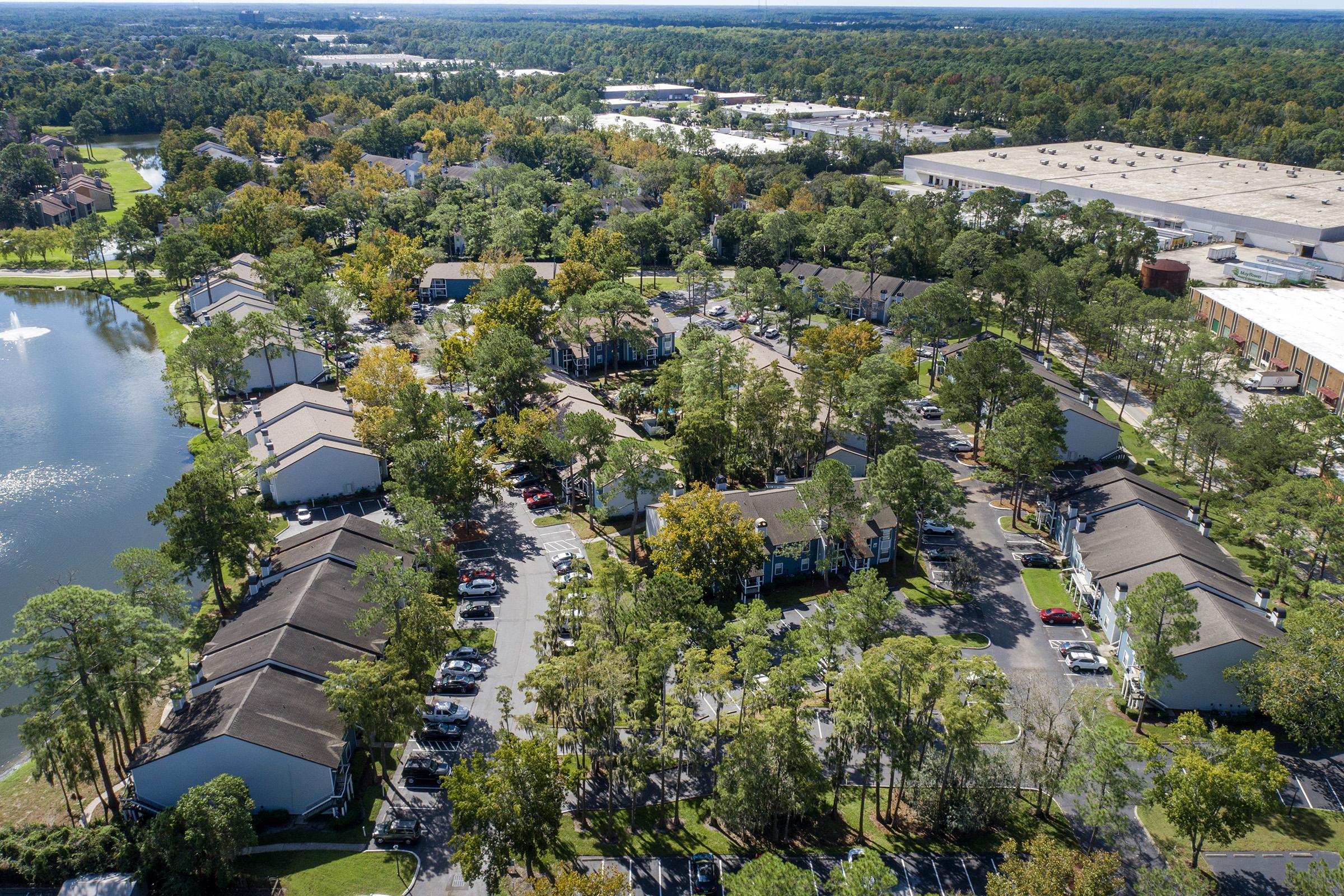 Aerial view of a residential area featuring houses arranged around a pond, surrounded by trees with autumn foliage. In the background, there are commercial buildings and a large parking lot. The scene conveys a serene neighborhood atmosphere.