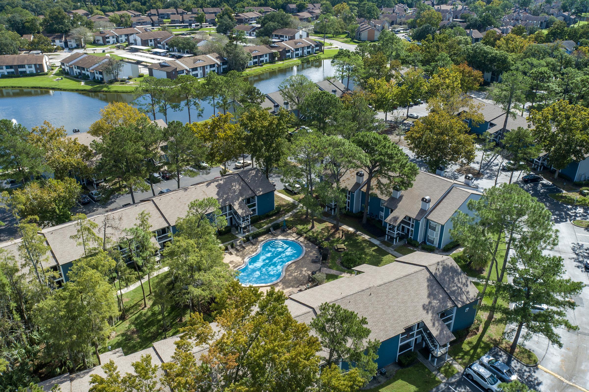 Aerial view of a residential community featuring multiple apartment buildings, a swimming pool surrounded by landscaping, and a serene pond in the background. The area is lush with trees and autumn foliage, highlighting a peaceful suburban atmosphere.
