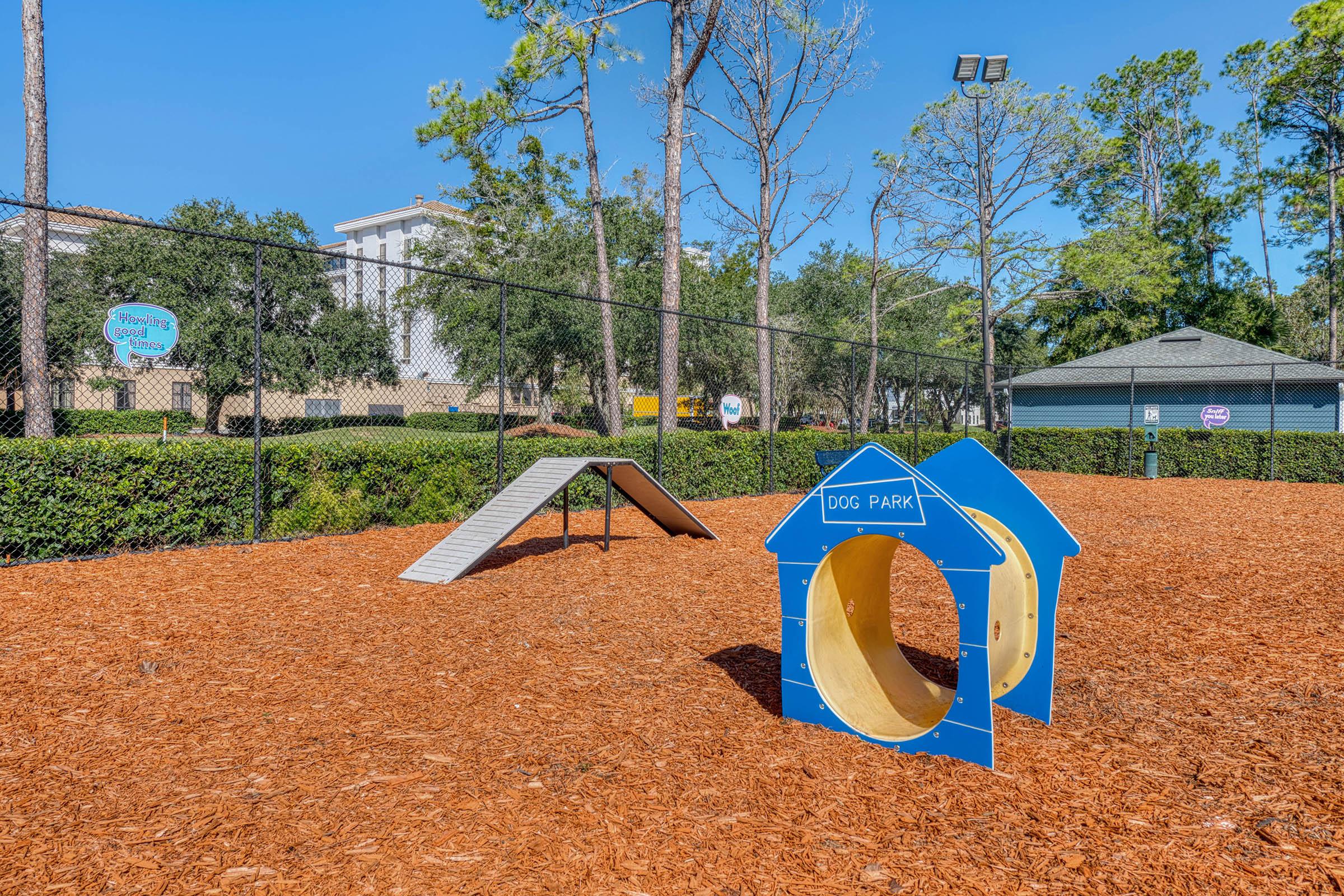 A dog park featuring a blue dog-shaped playhouse and an agility ramp on a bed of wood chips, surrounded by trees and a fenced area. A sign indicates the park's purpose, with nearby buildings and amenities visible in the background.