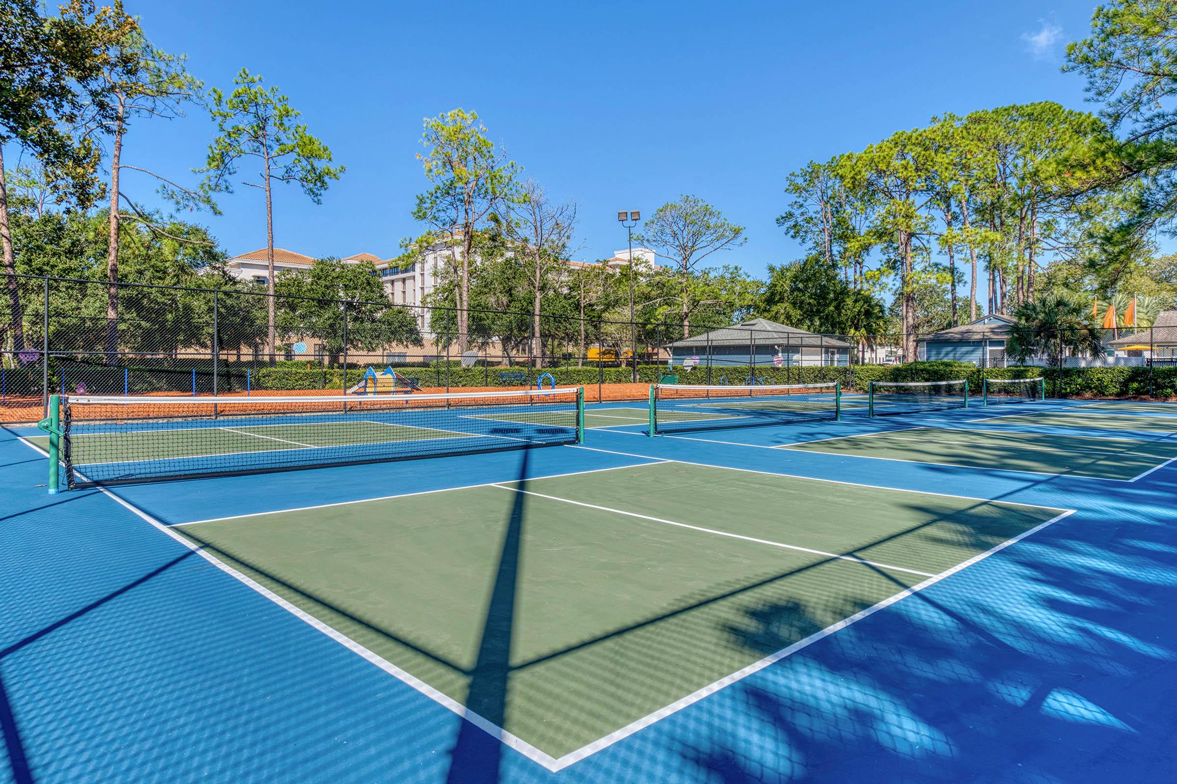 A view of outdoor tennis courts under a clear blue sky, surrounded by trees. The courts are freshly painted in blue and green, with nets in place. In the background, there are benches and a building partially visible, suggesting a recreational area.