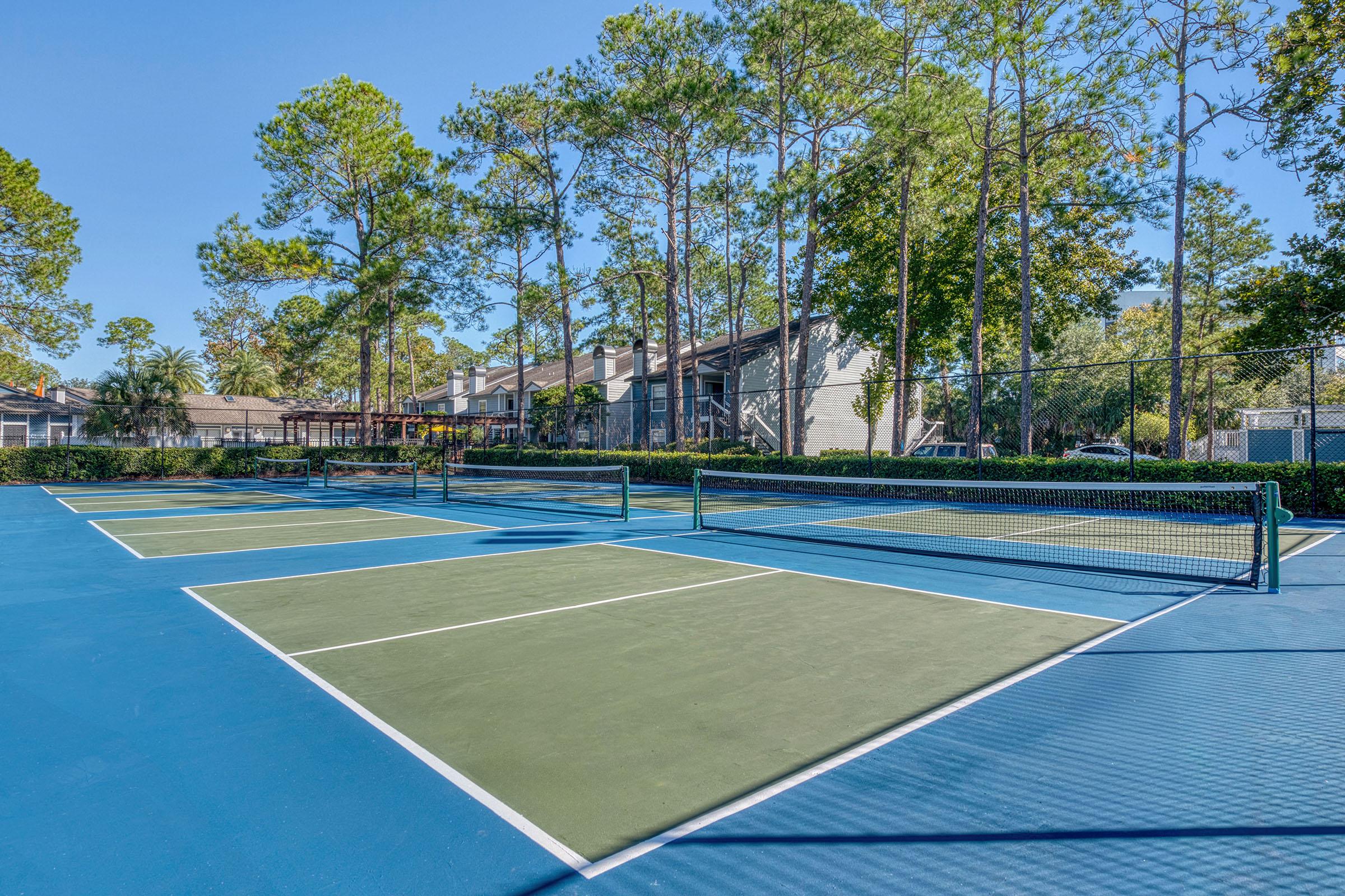 A bright outdoor tennis court surrounded by tall trees and residential buildings. The court features blue and green playing surfaces with two tennis nets set up. The clear sky adds to the serene atmosphere, making it a perfect setting for recreational play.