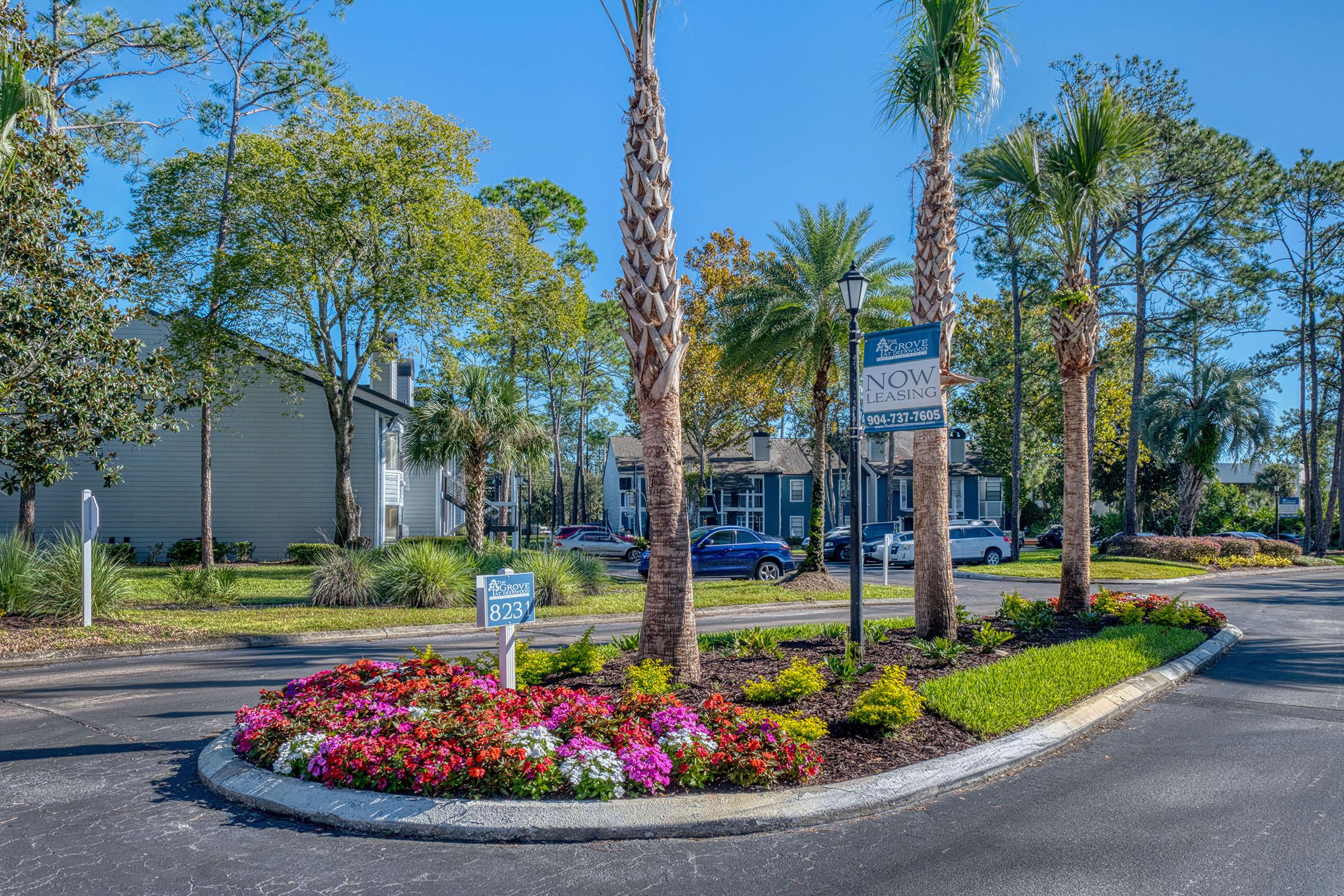 A landscaped entrance to an apartment complex featuring palm trees and colorful flowerbeds. A sign reads "Now Leasing" alongside the road, with vehicles parked nearby. The scene is set on a sunny day, surrounded by greenery and residential buildings.