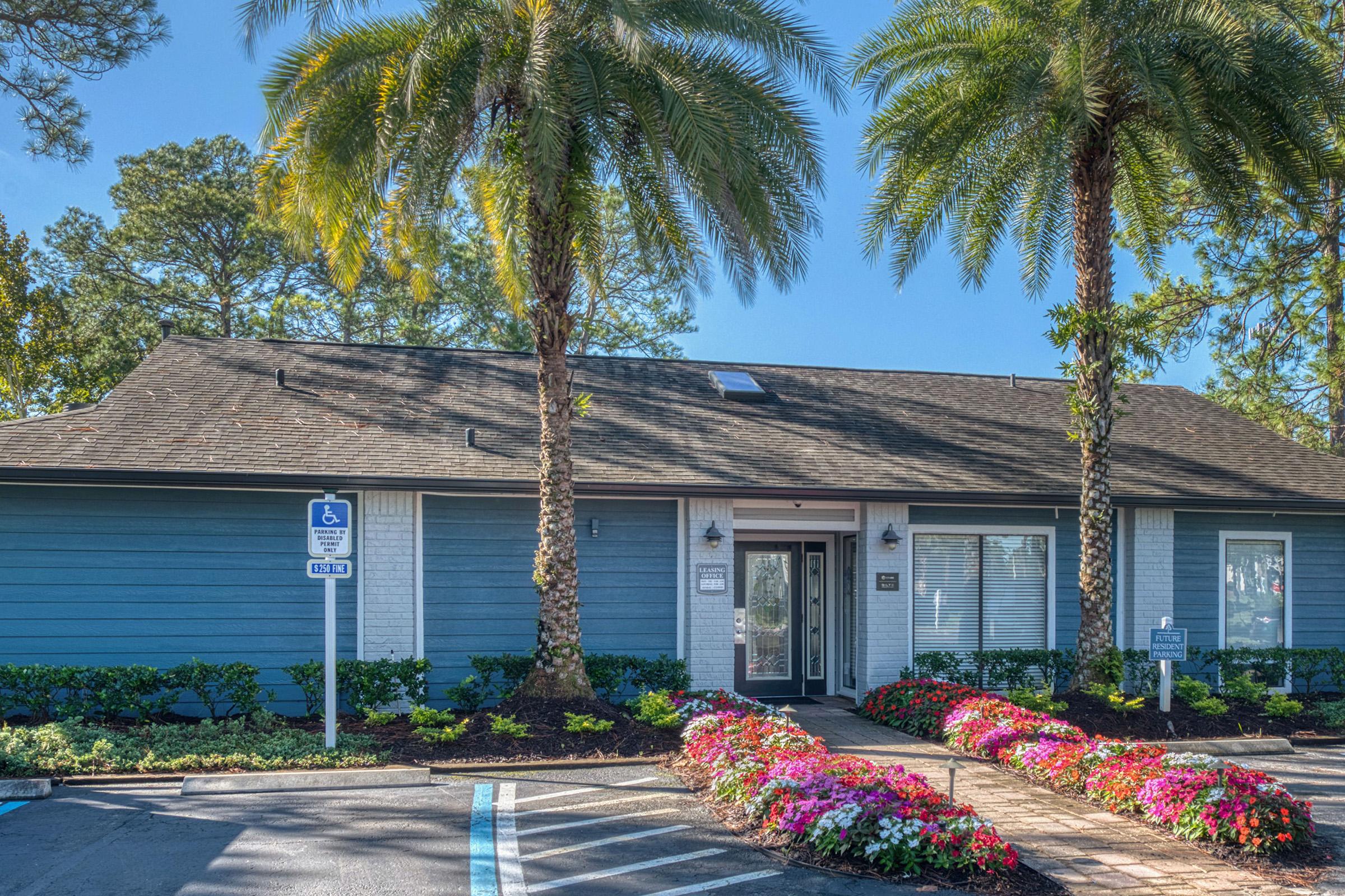 A welcoming building with a blue exterior and a gray roof, surrounded by palm trees. Colorful flower beds with vibrant blooms line the path leading to the entrance, which features large windows and elegant lighting. Accessible parking is available nearby, creating an inviting atmosphere.