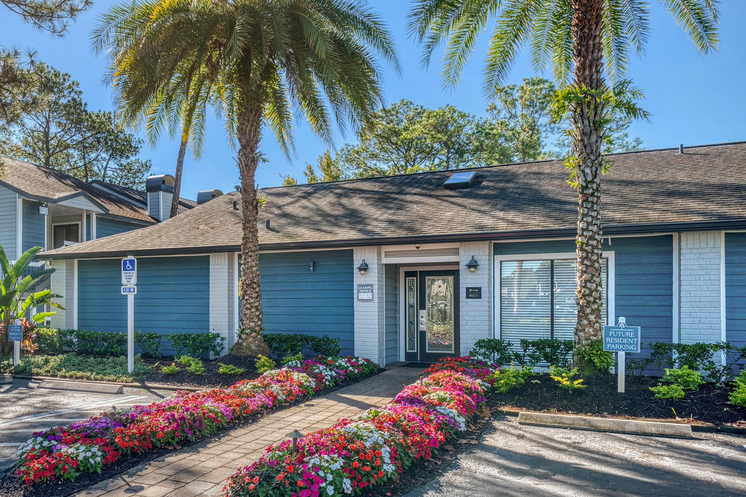 A well-maintained entrance of a residential building, featuring vibrant flowering plants and palm trees. The pathway is lined with colorful flowers, leading to a welcoming door. There are signs indicating parking and future management details. The building has a modern design with blue and white accents.
