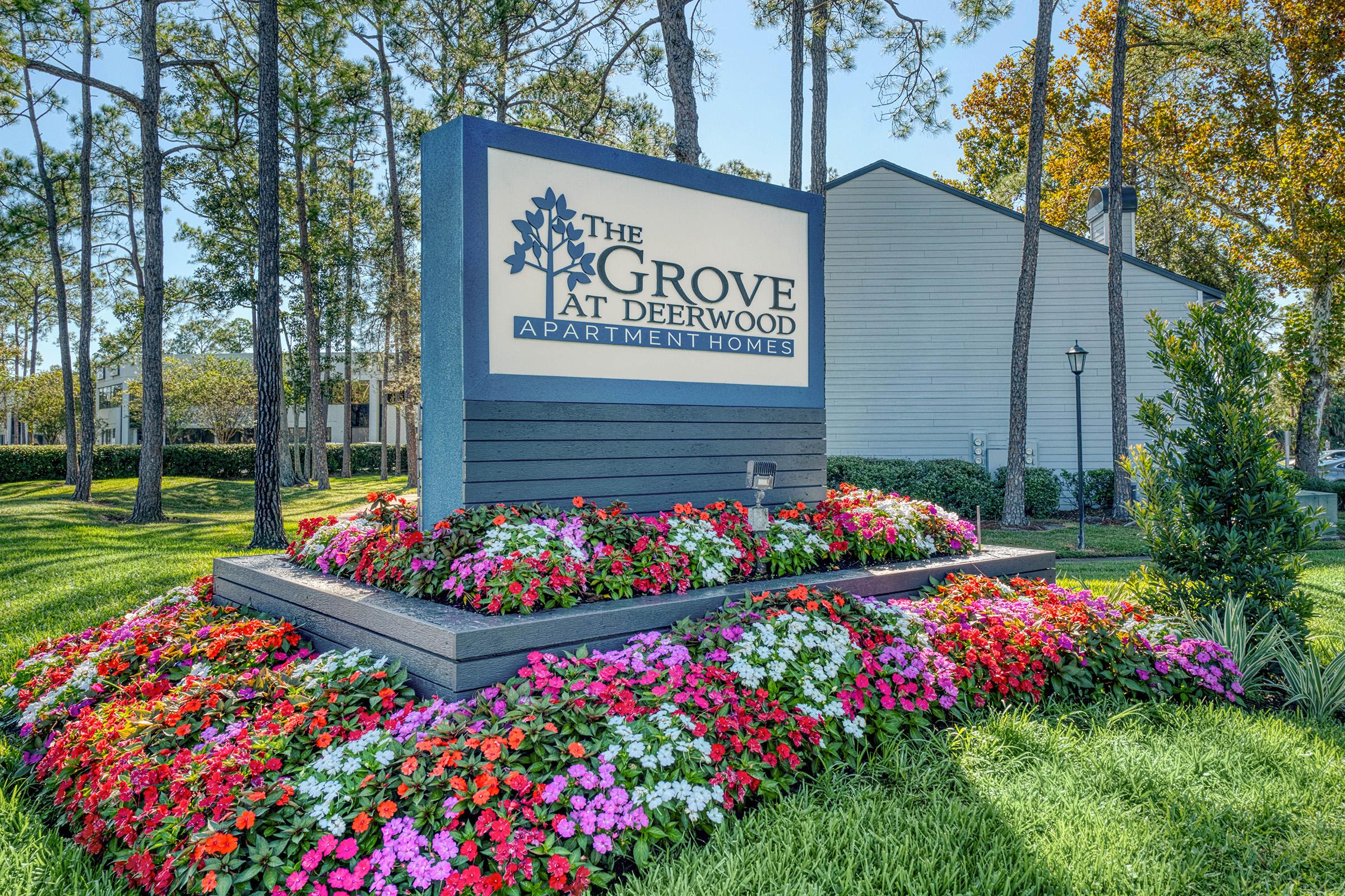 Sign for "The Grove at Deerwood Apartment Homes" surrounded by colorful flower beds and greenery, with tall trees in the background and a well-maintained landscape.