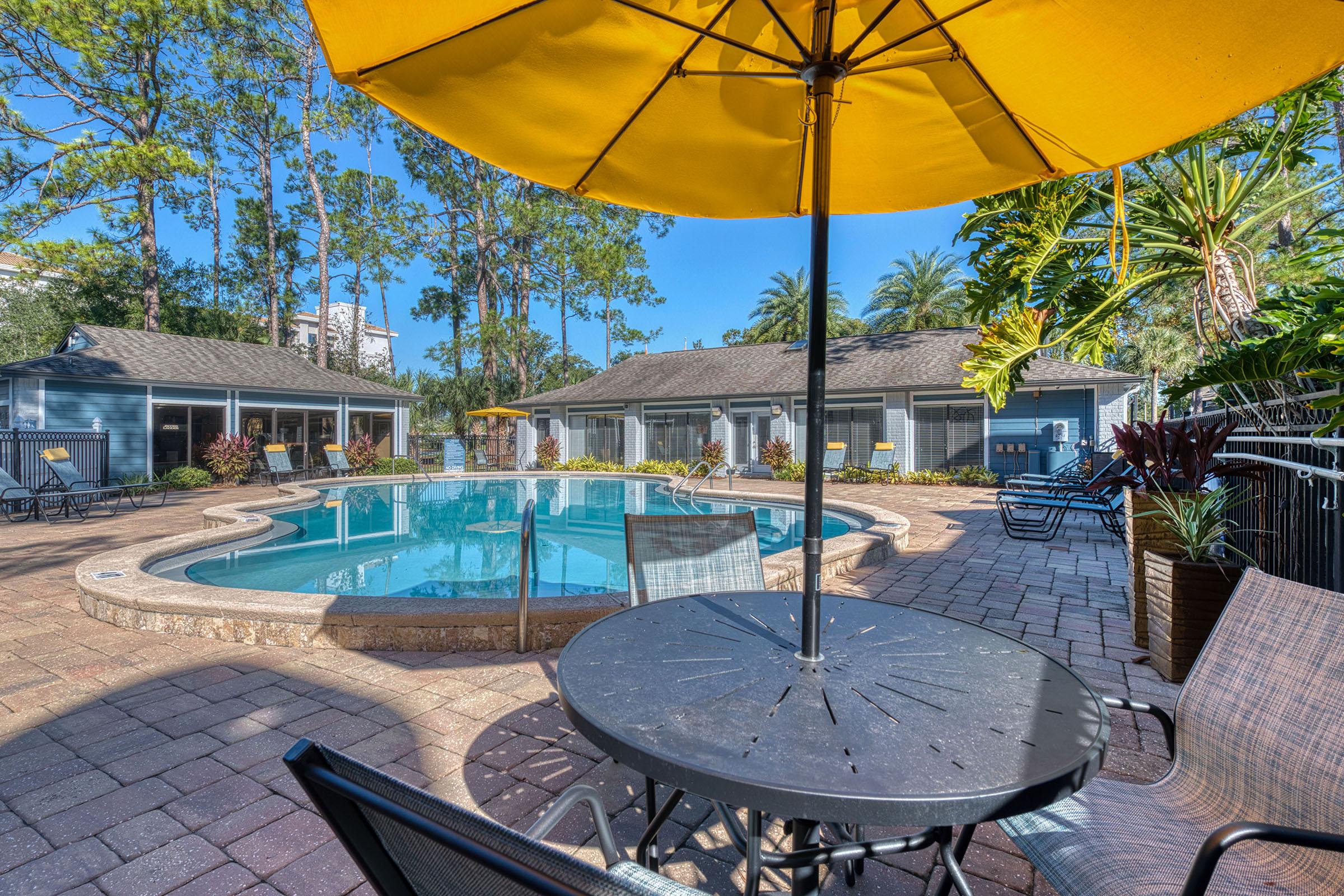 A sunny poolside area featuring a blue swimming pool, surrounded by lounge chairs and tropical plants. A yellow umbrella is positioned over a black table and chairs, adding a vibrant touch to the scene. In the background, there are buildings with screens, under a clear blue sky.