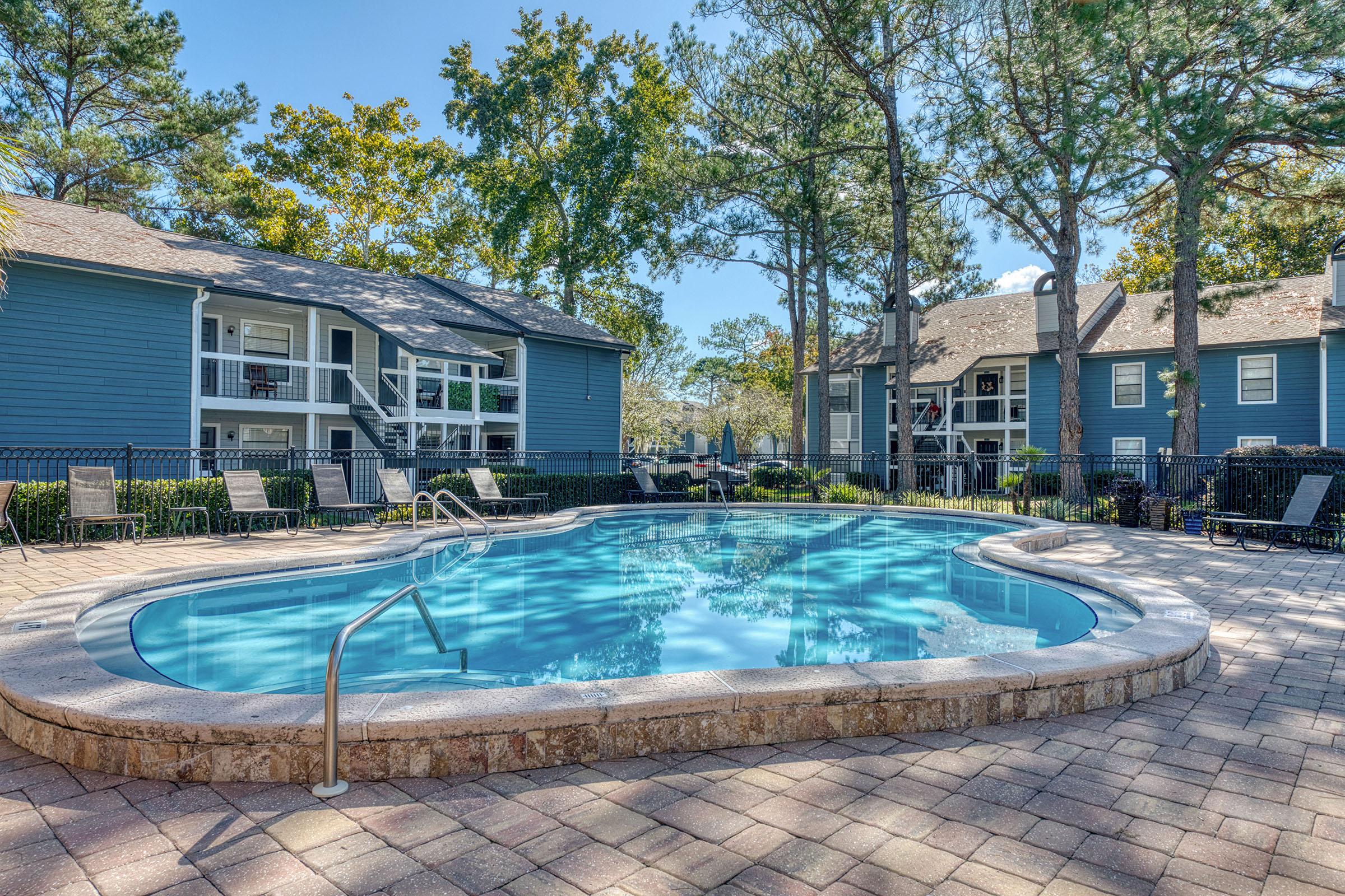 A serene outdoor swimming pool surrounded by greenery, with blue water reflecting the sky. Lounge chairs are positioned around the pool area. In the background, several two-story apartment buildings are visible, framed by tall trees and lush foliage, creating a peaceful residential atmosphere.