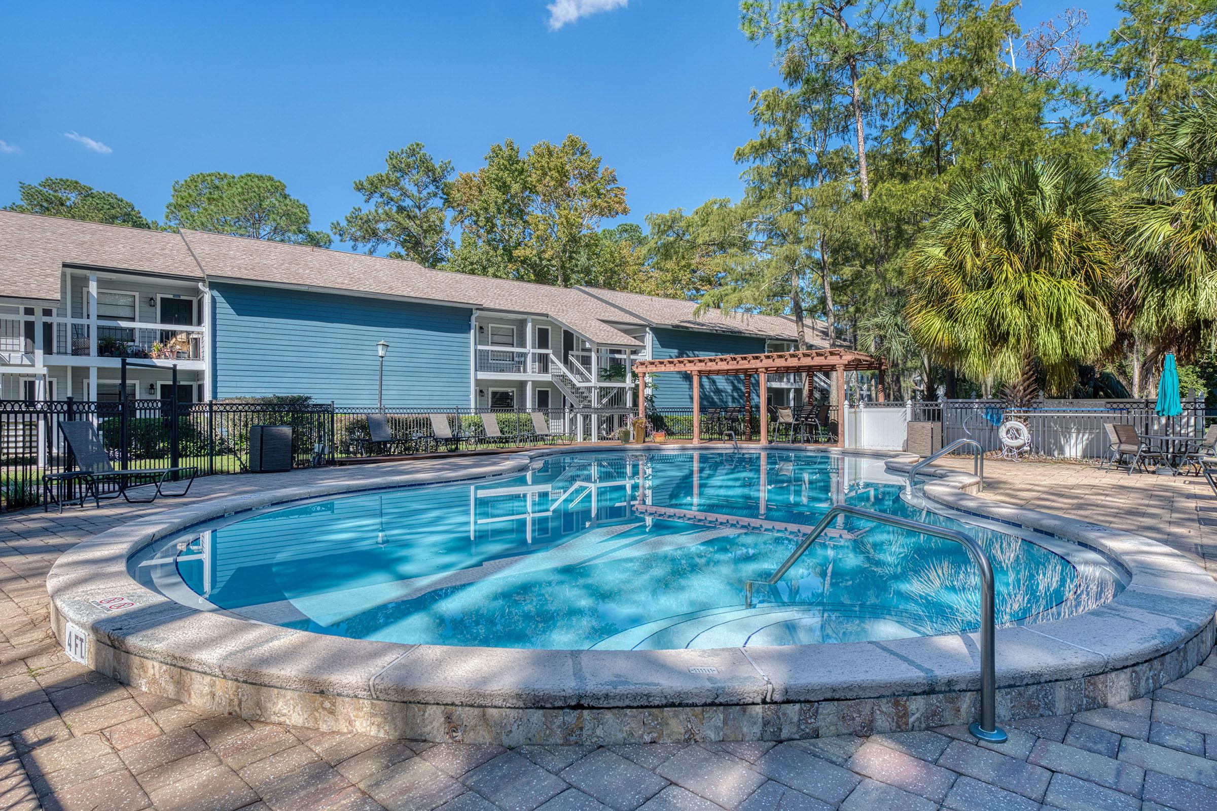A serene swimming pool surrounded by a stone patio, featuring a handrail for entry. Lush trees and greenery frame the scene, with comfortable seating around the pool area. Nearby, an apartment complex is visible in the background, highlighting a warm, inviting atmosphere. Clear blue skies set a relaxing vibe.