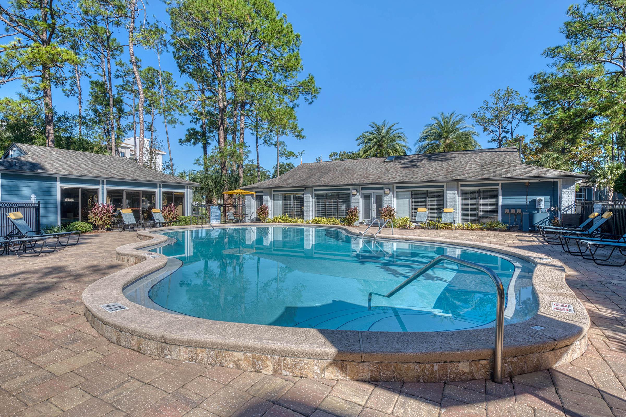 A clear swimming pool surrounded by a brick patio and lounge chairs. The area is shaded by tall trees, and there are two buildings with large windows in the background. Bright blue sky overhead complements the serene atmosphere of the pool area.