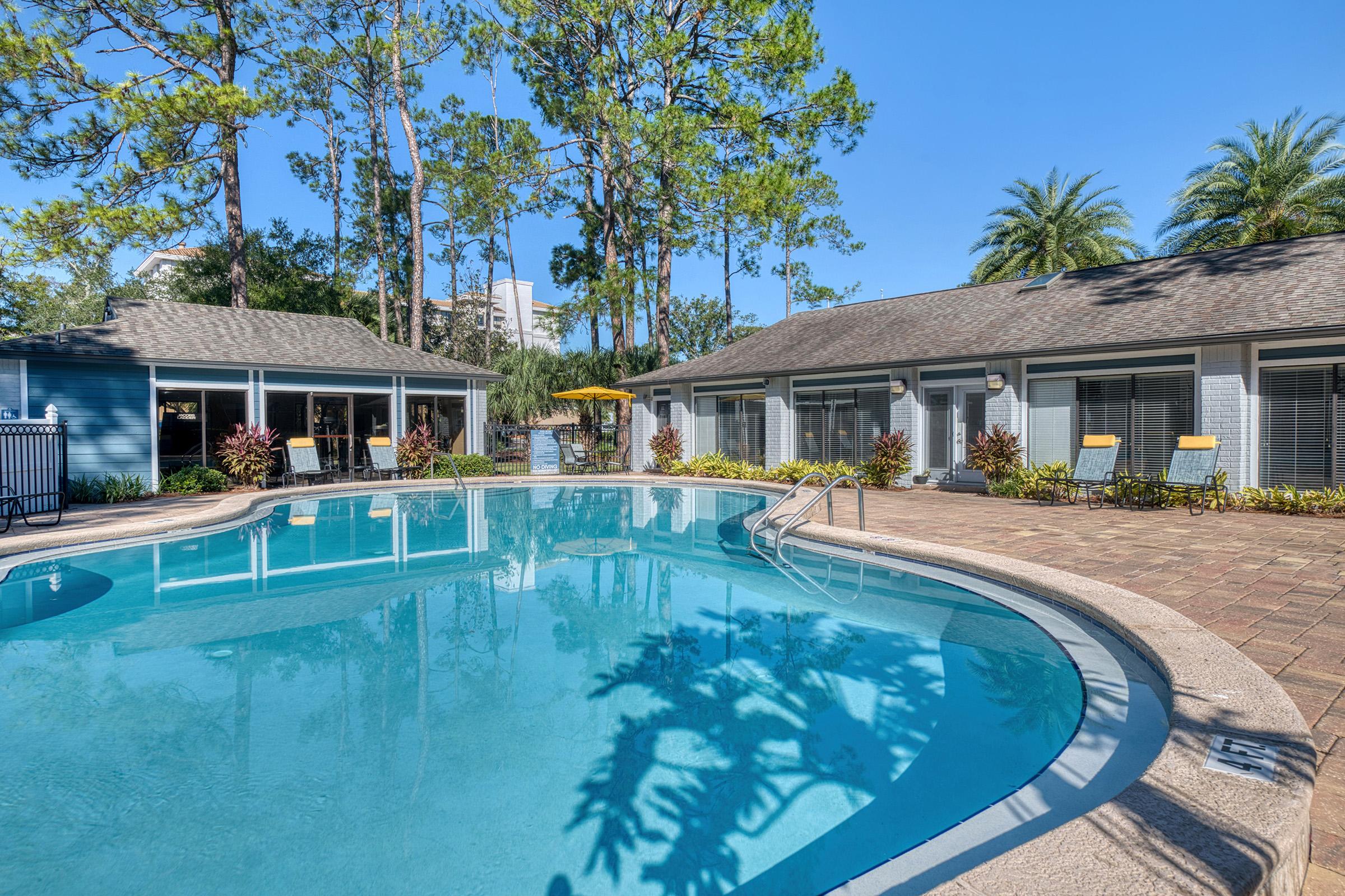 A serene pool area surrounded by lush greenery, featuring palm trees and tall pines. The water is clear, reflecting the blue sky. Two adjacent buildings with outdoor seating are visible near the pool, creating a relaxing ambiance perfect for leisure and relaxation.