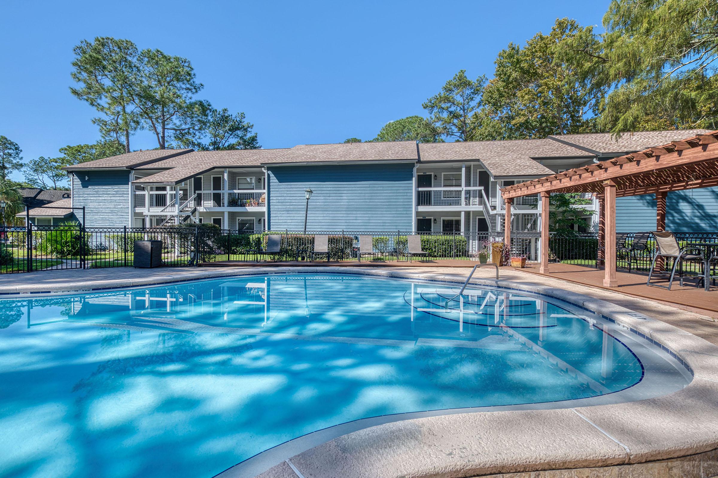 A clear blue swimming pool surrounded by a patio area with lounge chairs, adjacent to a residential building with multiple units. Lush trees can be seen in the background, and a pergola with seating is visible on one side of the pool, creating a relaxing outdoor atmosphere.