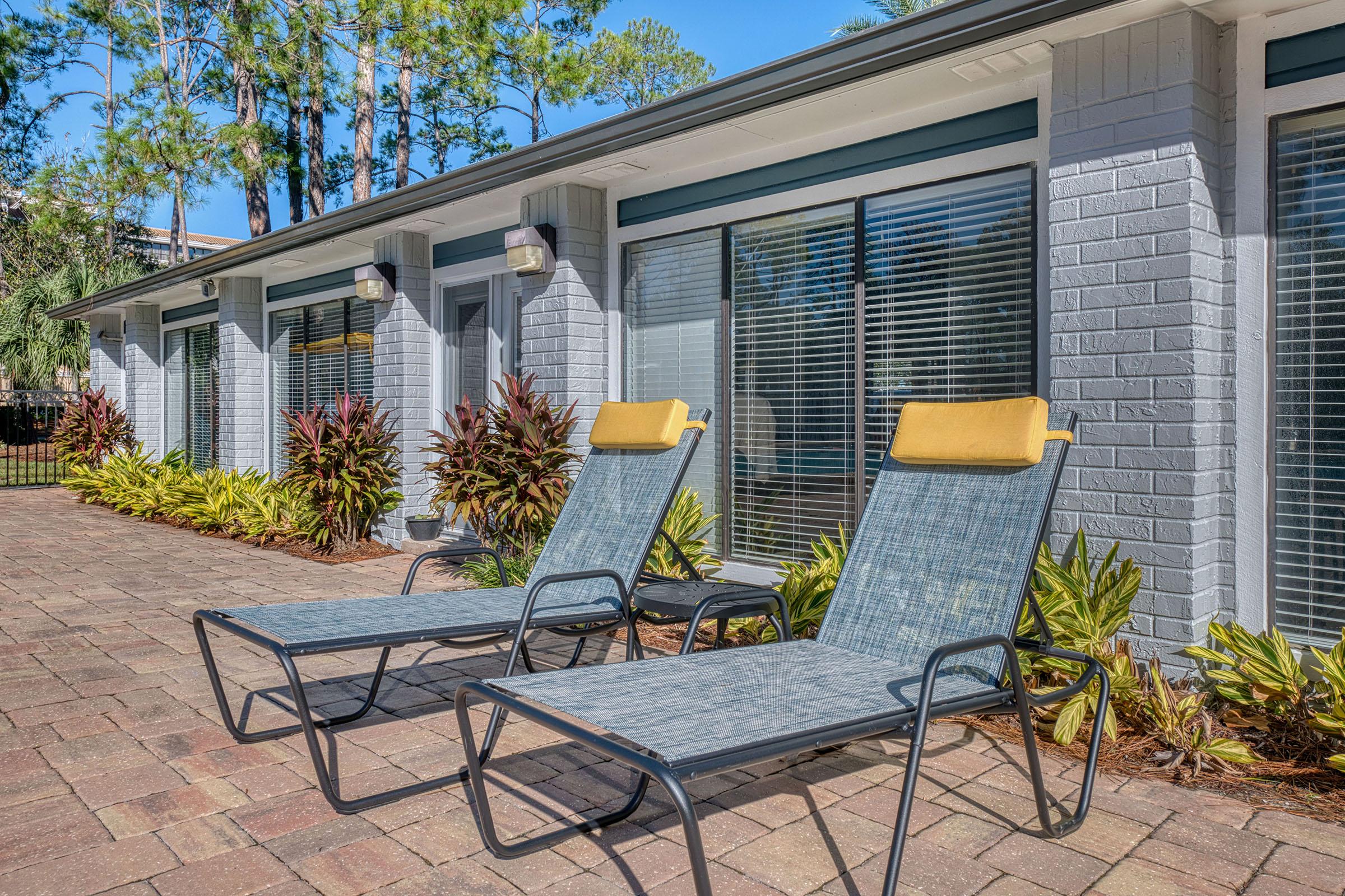 Two modern lounge chairs with yellow headrests are placed on a brick patio in front of a building. The area features vibrant green plants and trees. Large windows with horizontal blinds reflect the sunlight, creating a welcoming outdoor space for relaxation.