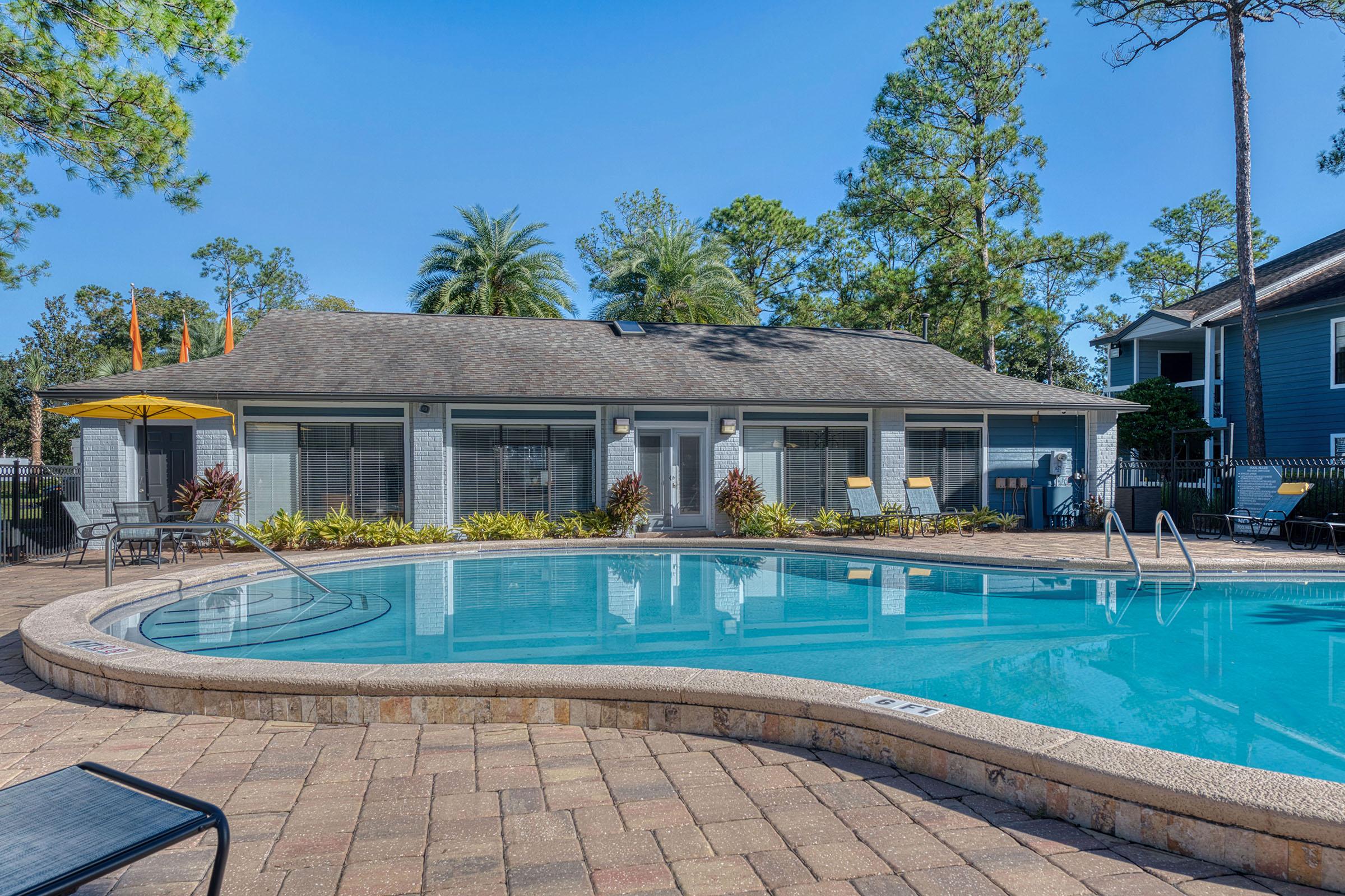 A serene swimming pool area surrounded by lush greenery and tall trees. The pool features a gentle curve, with a shallow entrance and lounge chairs nearby. A building with large windows sits in the background, providing a relaxing atmosphere under a clear blue sky. Perfect for leisure and relaxation.