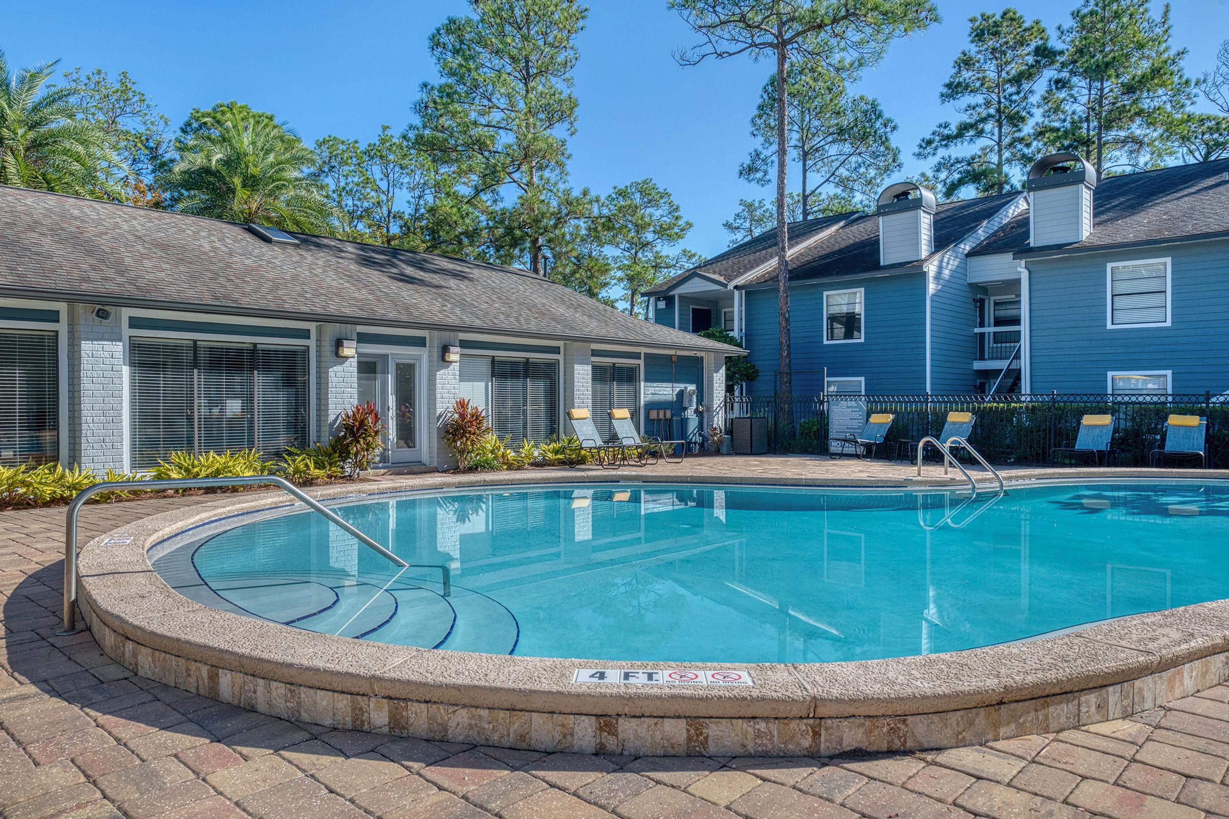 A serene swimming pool surrounded by lounge chairs and lush greenery. The pool area features a curved edge and is bordered by a brick patio. Nearby, there are two residential buildings painted in blue, with large windows and a peaceful atmosphere under a clear sky.