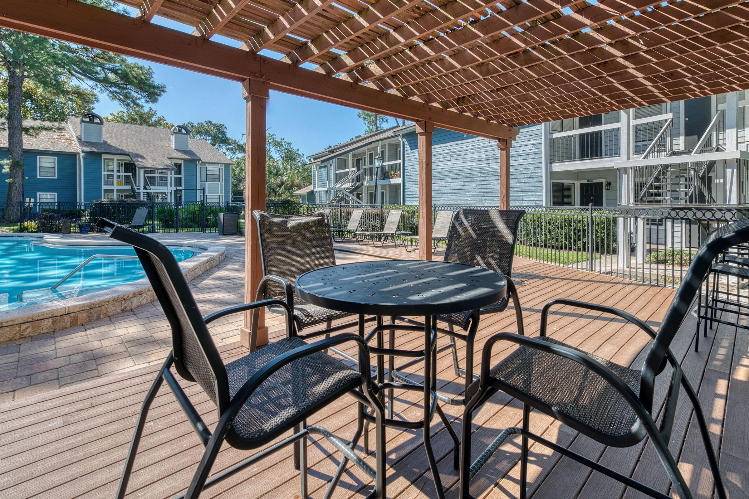 A shaded outdoor seating area with a table and four chairs overlooks a swimming pool. Surrounding the pool are lounge chairs and apartments in the background, with trees providing a pleasant, green backdrop under a clear blue sky.