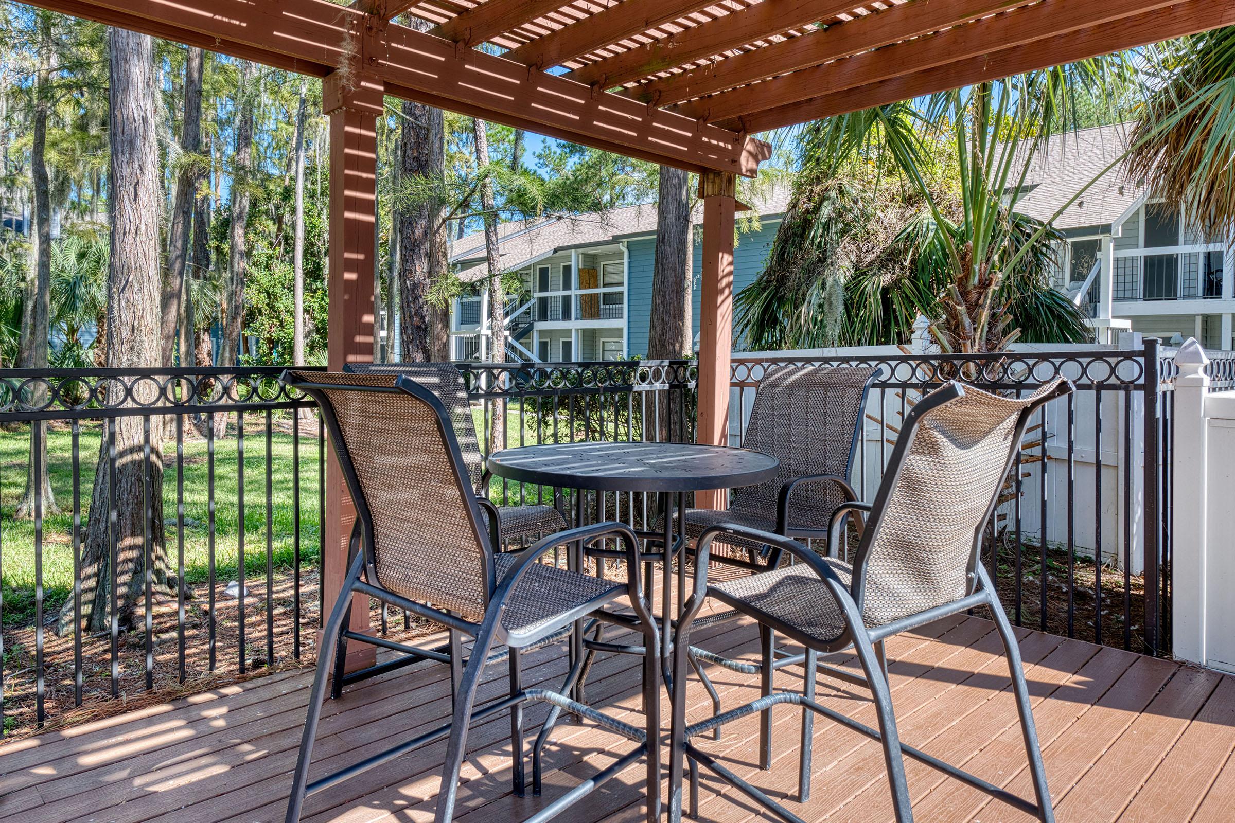 A cozy outdoor patio featuring a small round black table and four wicker chairs, shaded by a wooden pergola. The background includes lush green trees and nearby residential buildings, creating a serene and inviting atmosphere for relaxation or dining.