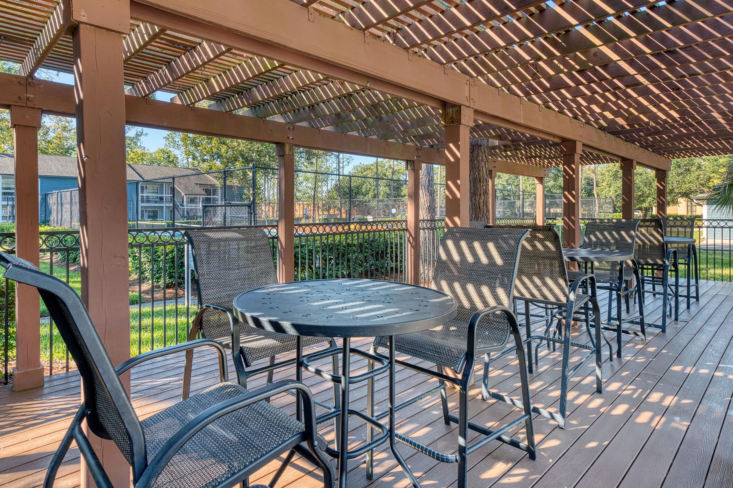 A spacious outdoor patio with black metal furniture, including round tables and chairs, under a wooden pergola. Soft sunlight filters through the slats, casting shadows on the deck. The surrounding greenery and a fence create a serene atmosphere, perfect for relaxation or social gatherings.