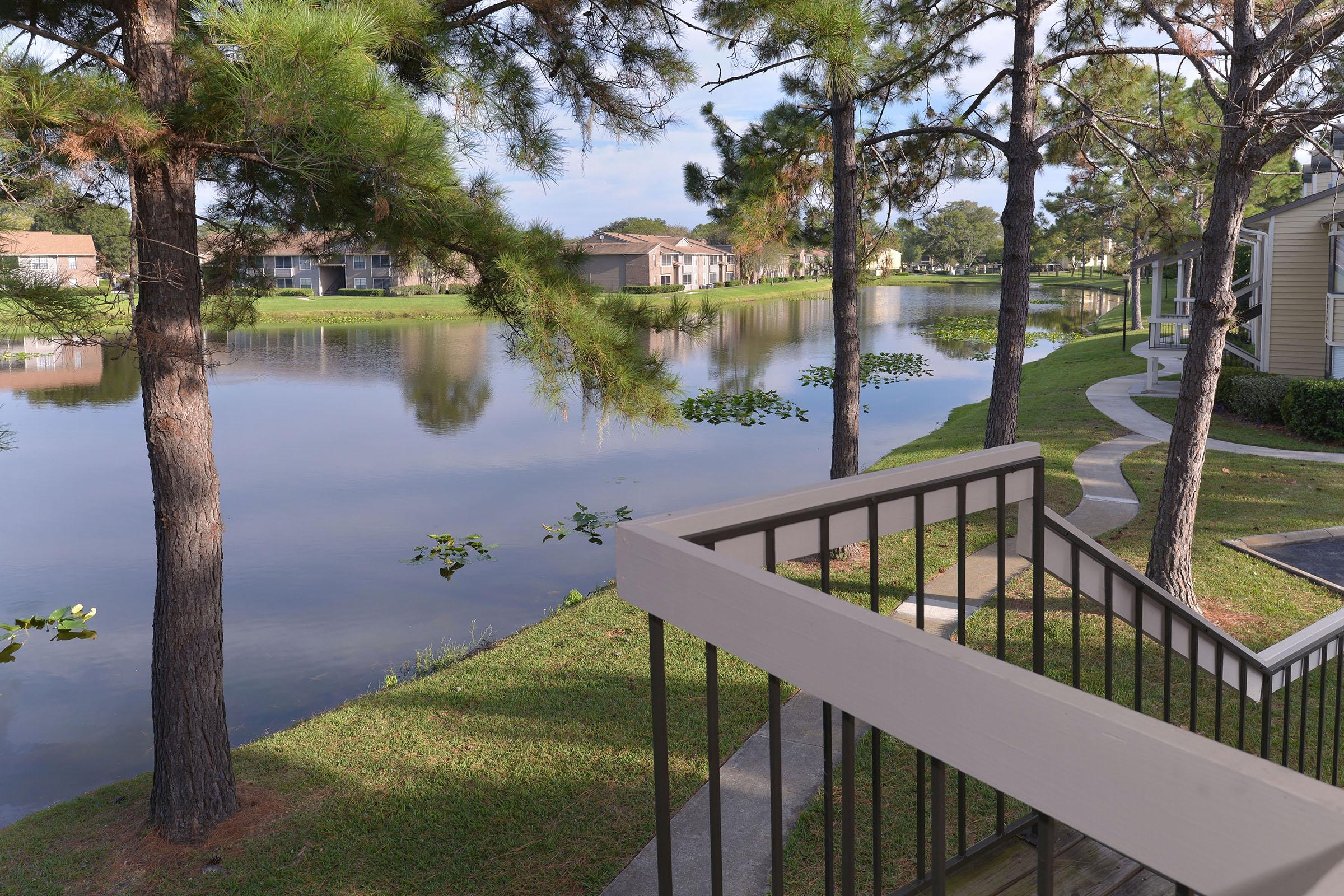 View of a serene lakeside scene featuring a calm body of water surrounded by lush greenery and tall trees. There's a well-maintained pathway leading to the water's edge, with several residential buildings visible in the background, reflecting a peaceful suburban atmosphere.