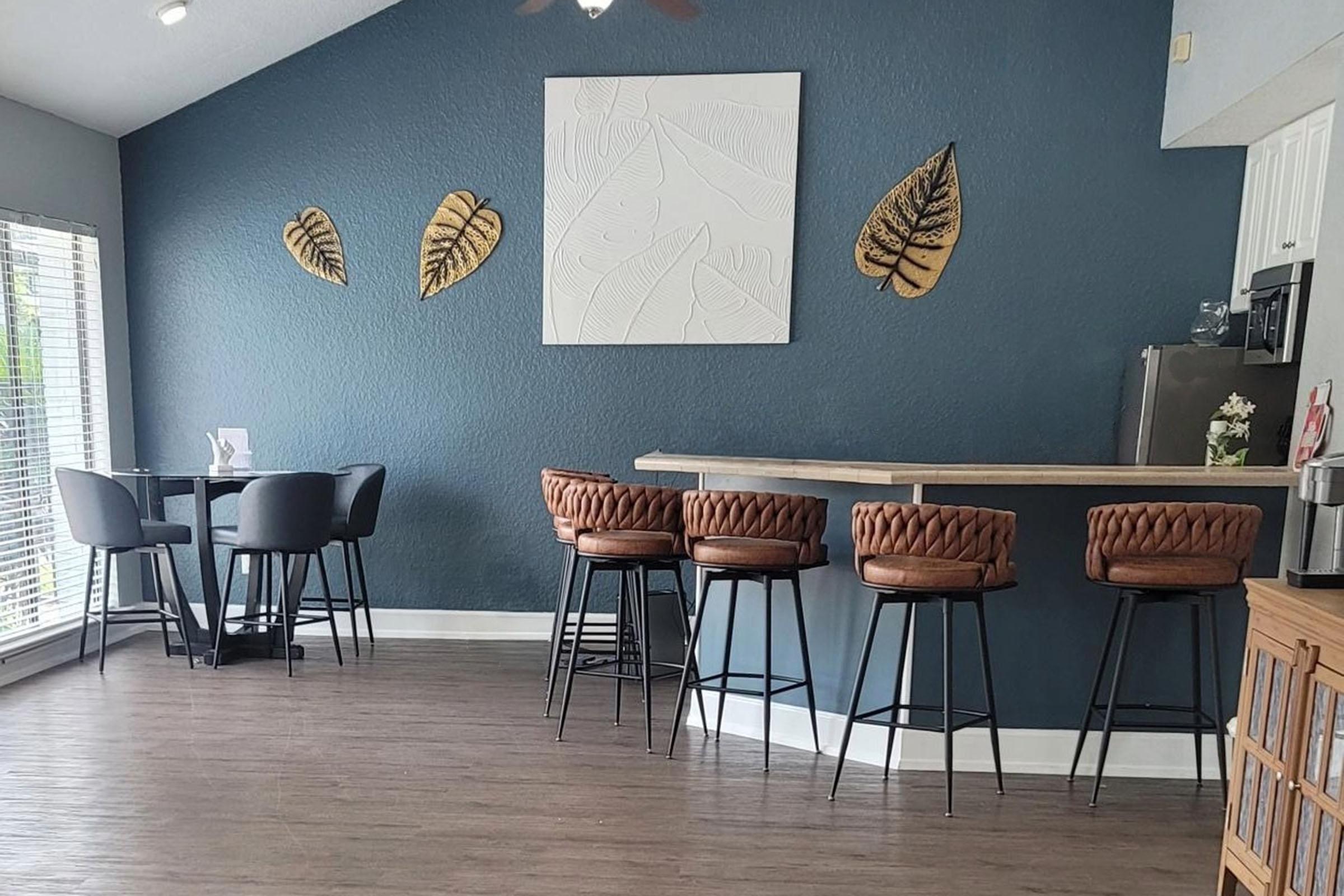 A modern interior featuring a light blue wall adorned with decorative leaf art, a round dining table with black chairs, and a sleek bar area with brown cushioned stools. Large windows allow natural light to brighten the space, creating a cozy and inviting atmosphere.