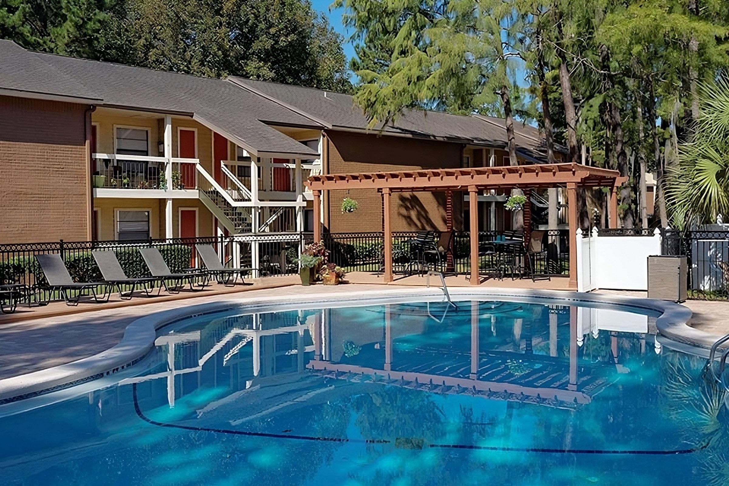 A clear swimming pool surrounded by lounge chairs, with a pergola and tables nearby. In the background, several apartment buildings are visible, set against a backdrop of trees. The scene is bright and inviting, suggesting a relaxed outdoor atmosphere.