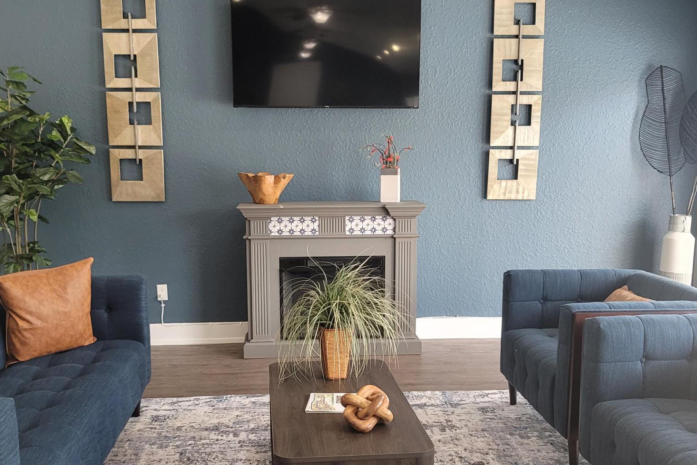 A modern living room featuring two blue sofas with brown cushions, a coffee table with a decorative plant and a book, and a faux fireplace. The walls are painted a dark blue, adorned with gold-framed artwork. A flat-screen TV is mounted above the fireplace, and a green potted plant is beside the seating area.
