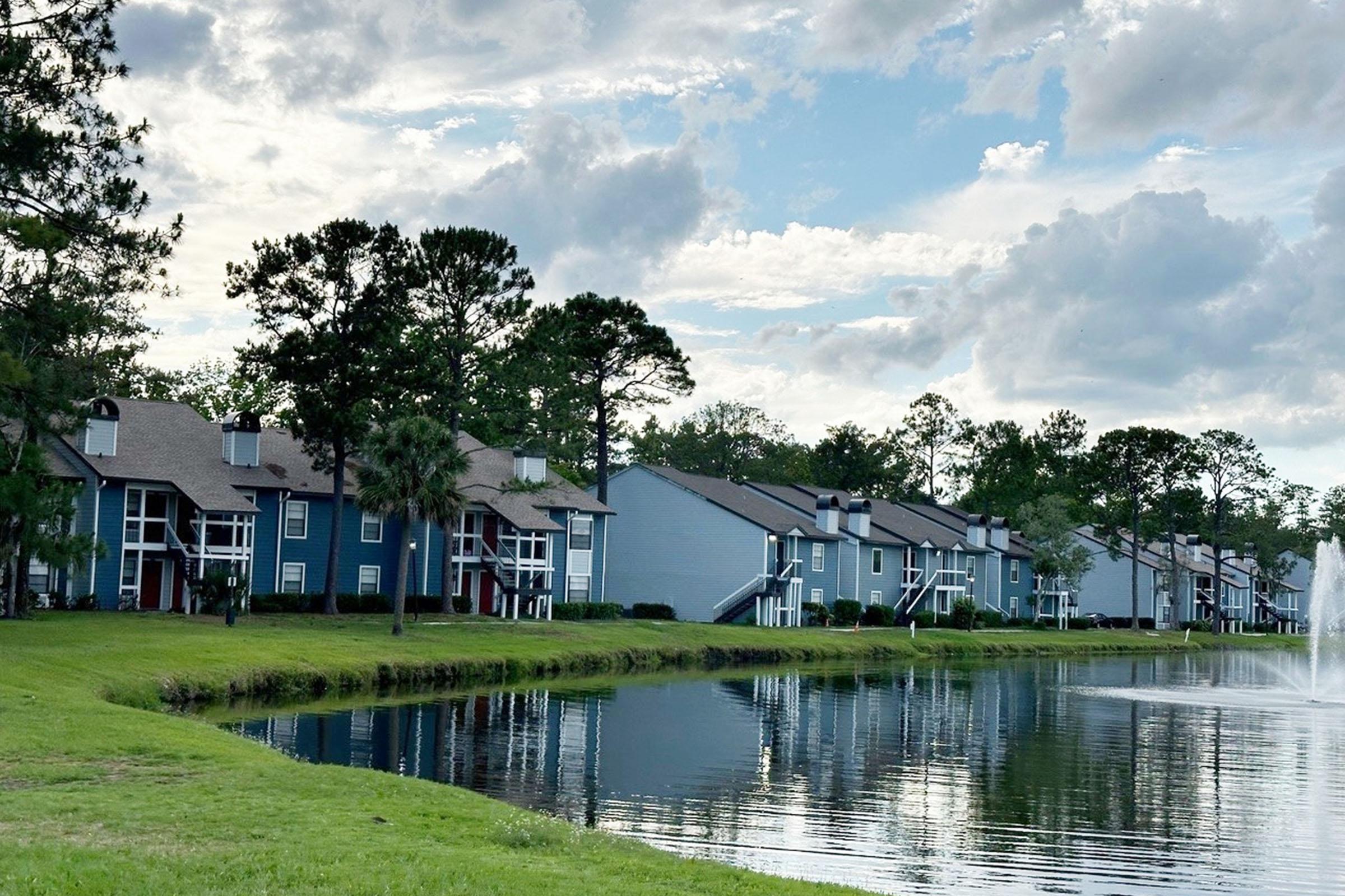 Row of residential buildings in shades of blue and gray beside a calm pond. Lush green grass and trees line the water's edge, with a fountain spraying gently in the background. The sky is partly cloudy, creating a serene and peaceful atmosphere.