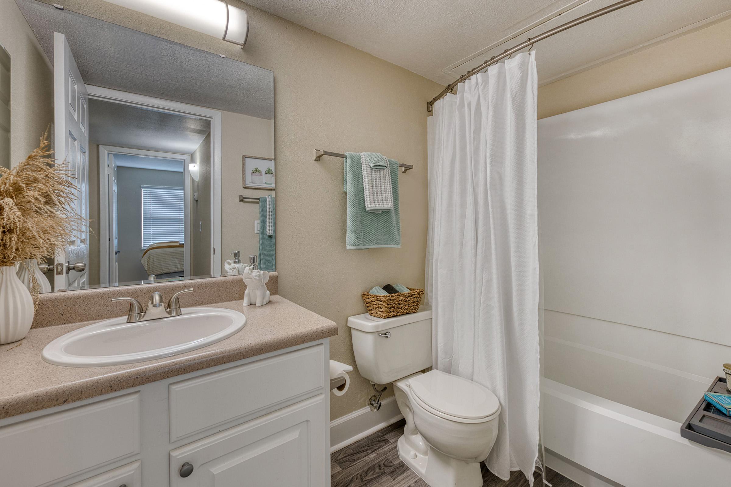 A clean and modern bathroom features a white vanity with a sink, a large mirror, a shower with a white curtain, a toilet, and a basket on the toilet tank. The decor includes a vase with dried flowers and a towel rack with a soft-colored towel. There is a glimpse of a bedroom through the open door.