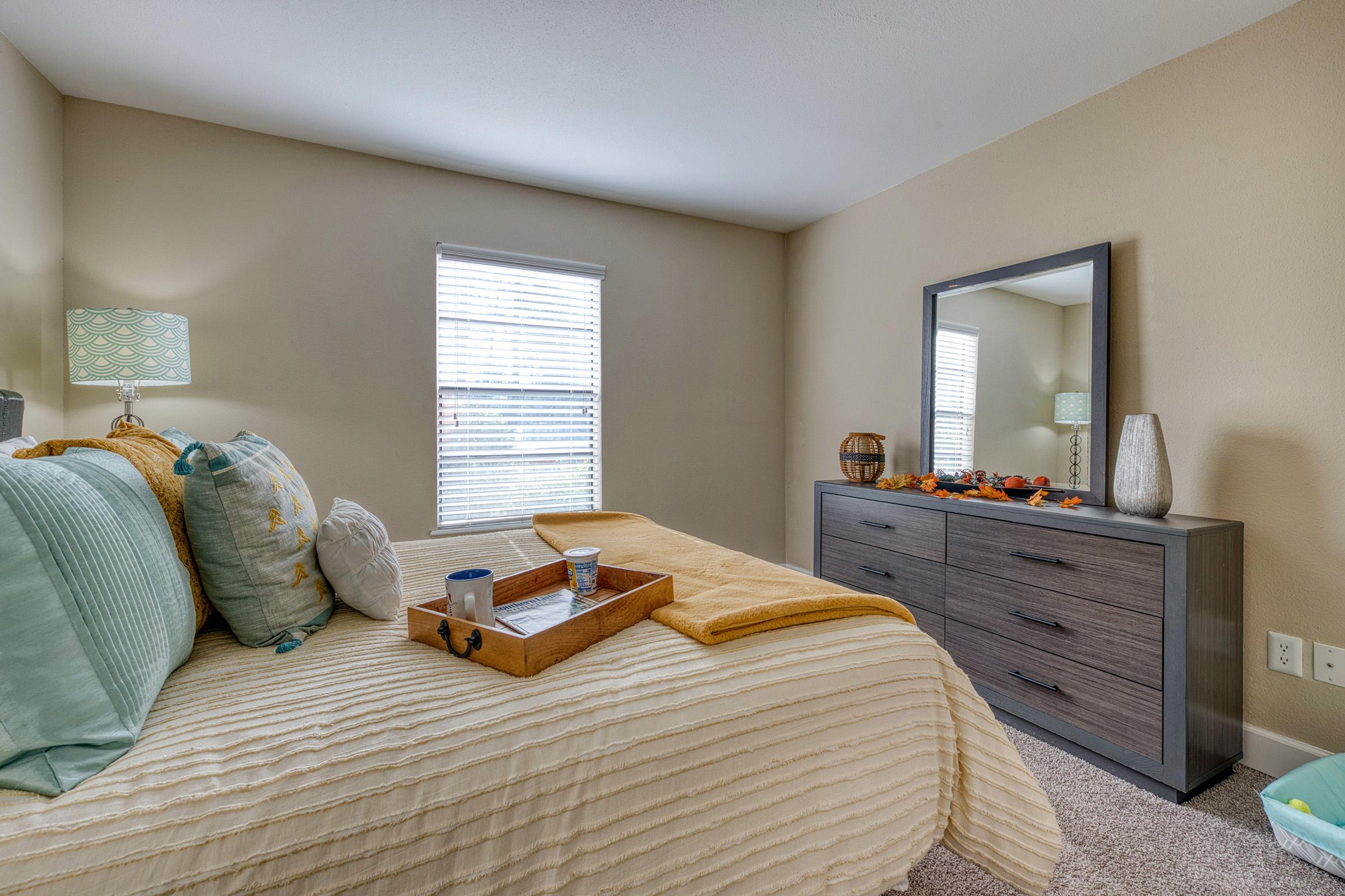 A cozy bedroom featuring a neatly made bed with decorative pillows and a tray. A large mirror sits on a dresser, and a soft, warm blanket is draped over the bed. Natural light streams through the window with blinds, and autumn decorations are placed on the dresser.